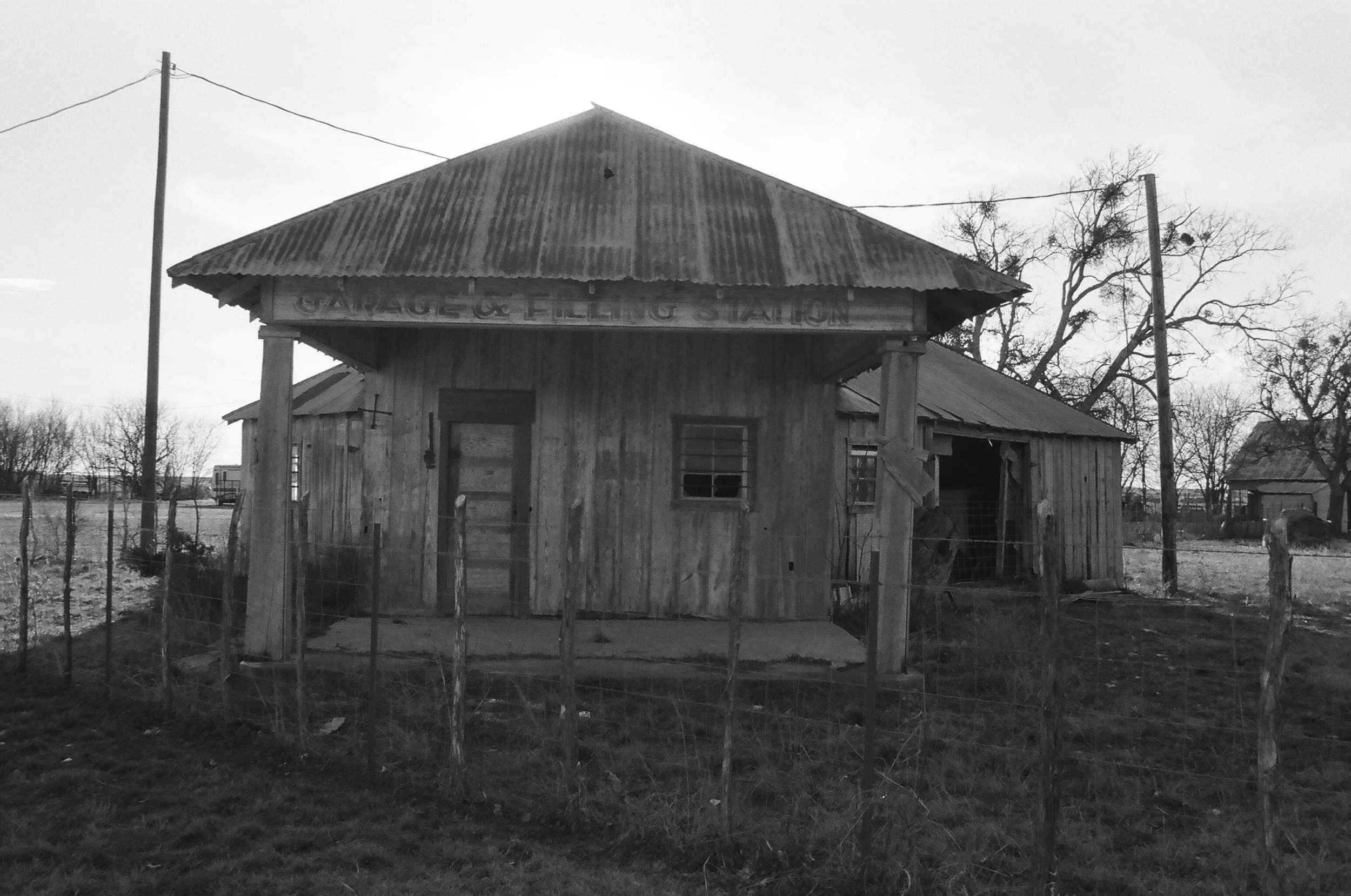 Old wooden garage with a corrugated metal roof, weathered wood walls, and a sign reading 'Garage & Filling Station' on the front, surrounded by a wire fence with trees and houses in the background.