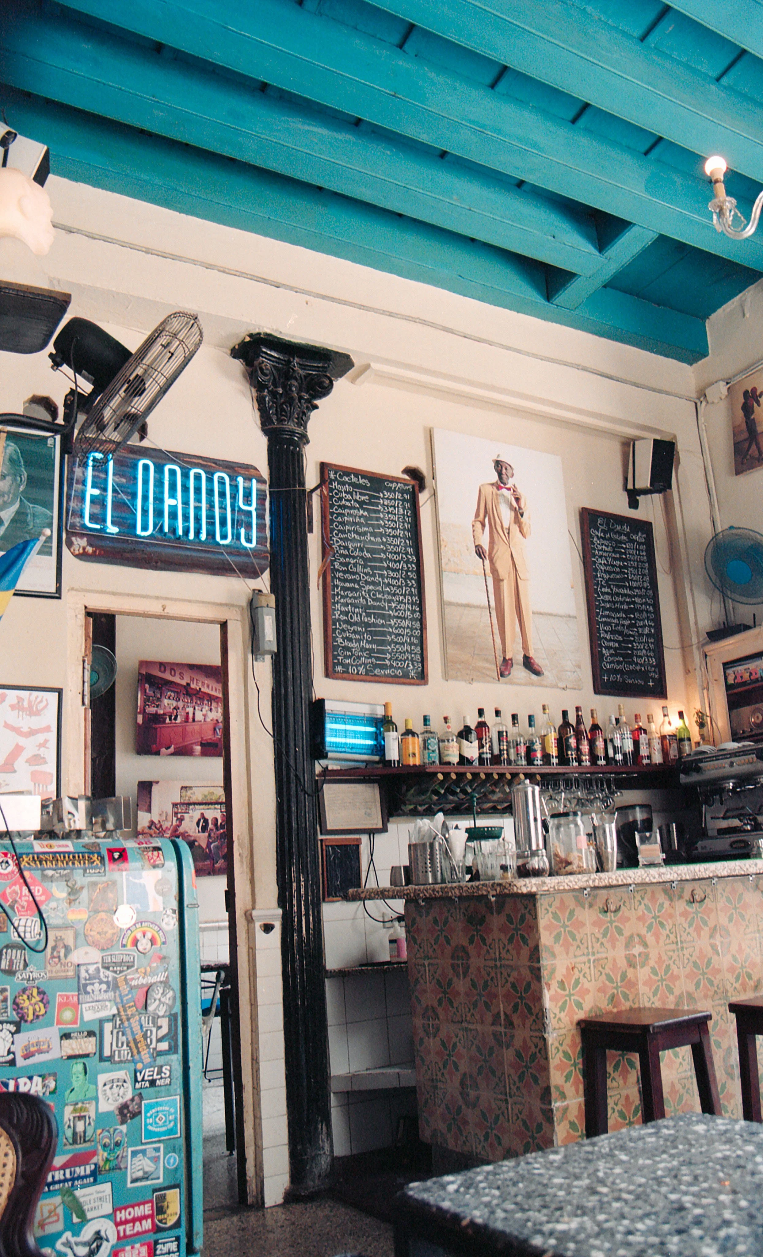 Interior of a bar or cafe with colorful tiled counter, shelves of bottles, promotional posters, neon sign that says 'EL AMOS', and a large photograph of a man in a light-colored suit and hat.