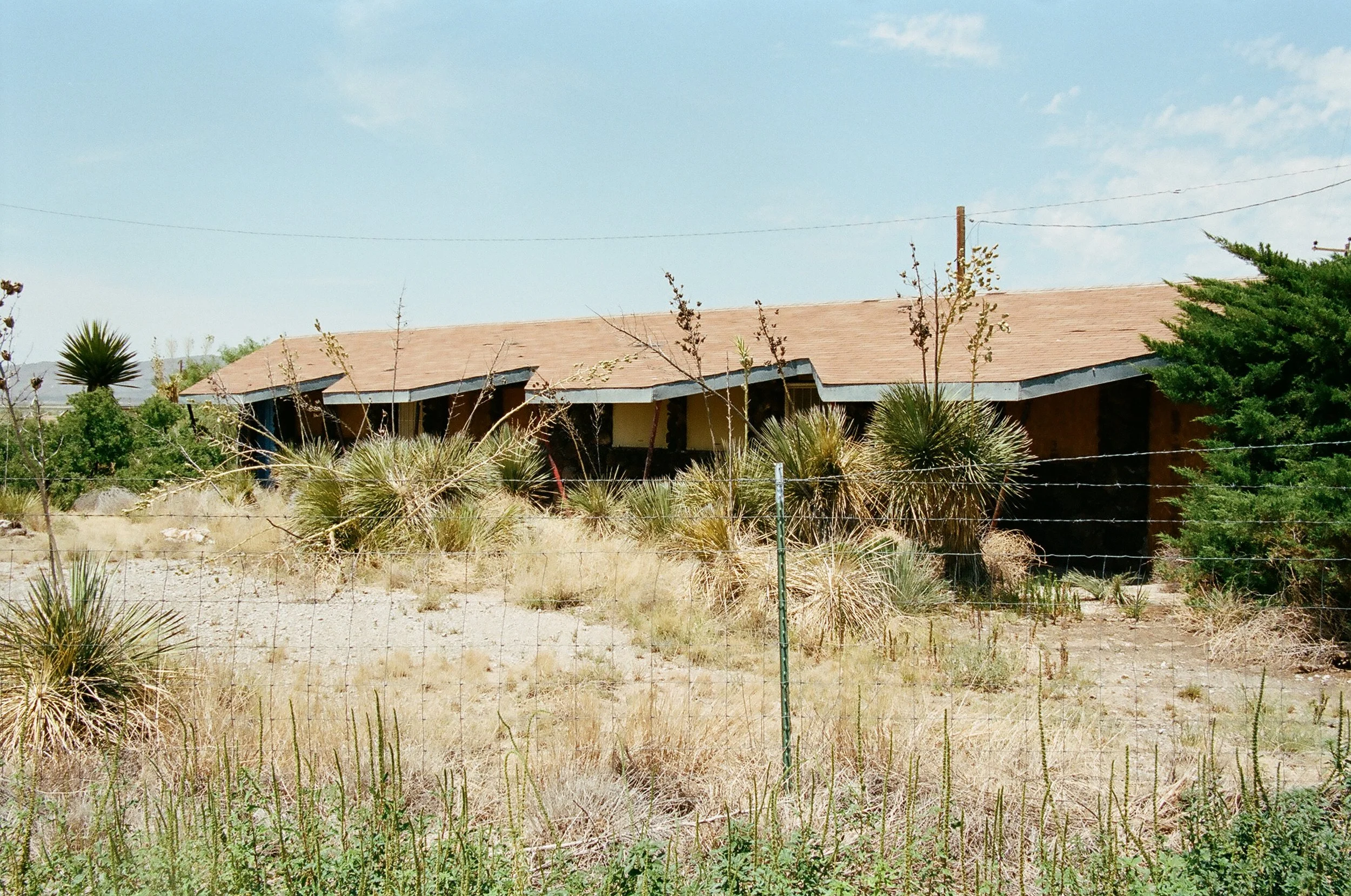 An abandoned house in a desert landscape with overgrown desert plants and a barbed wire fence in the foreground.