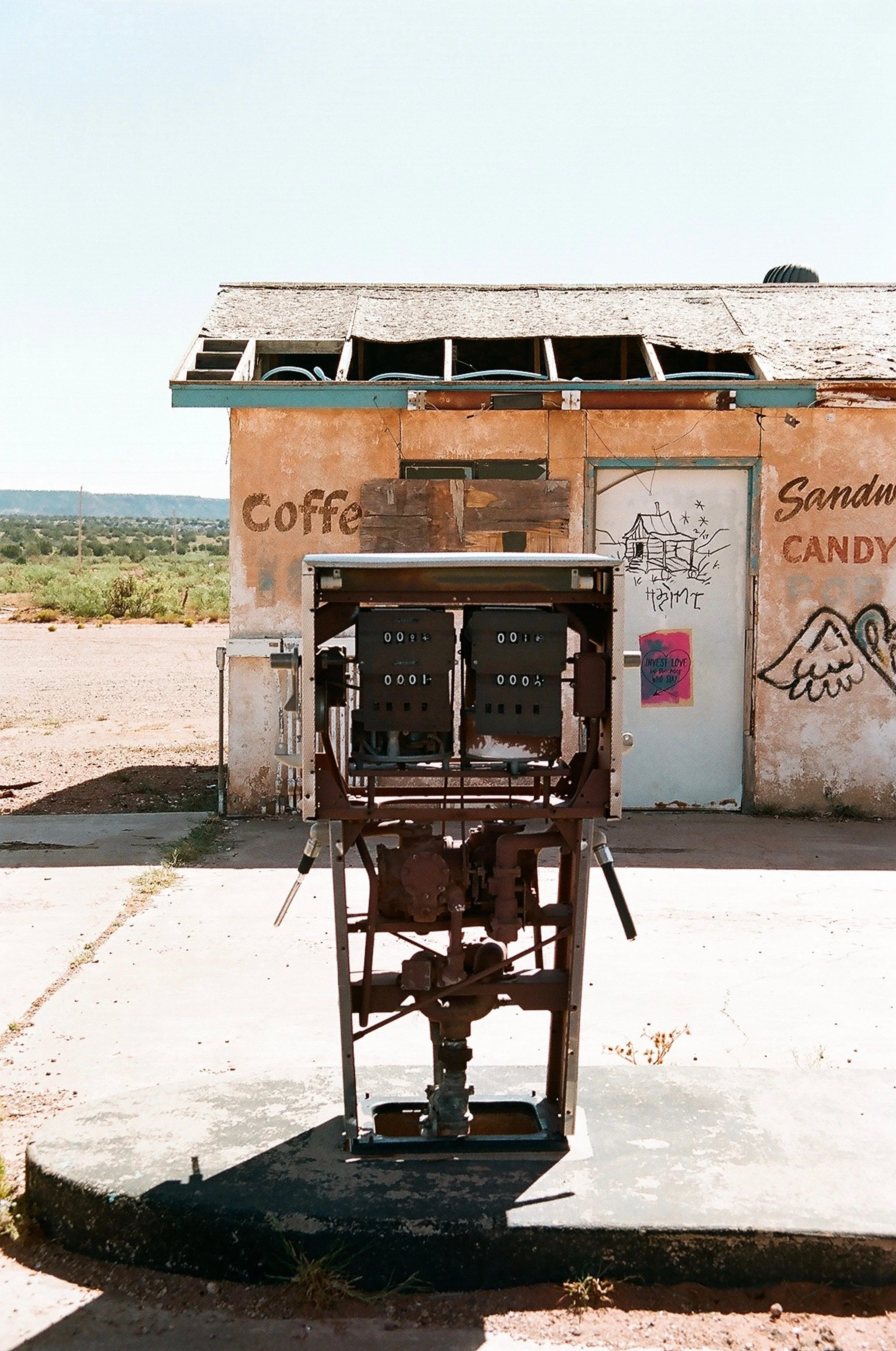 Empty, weathered small building with graffiti and signs, with an old mechanical device in front, in a desert landscape.