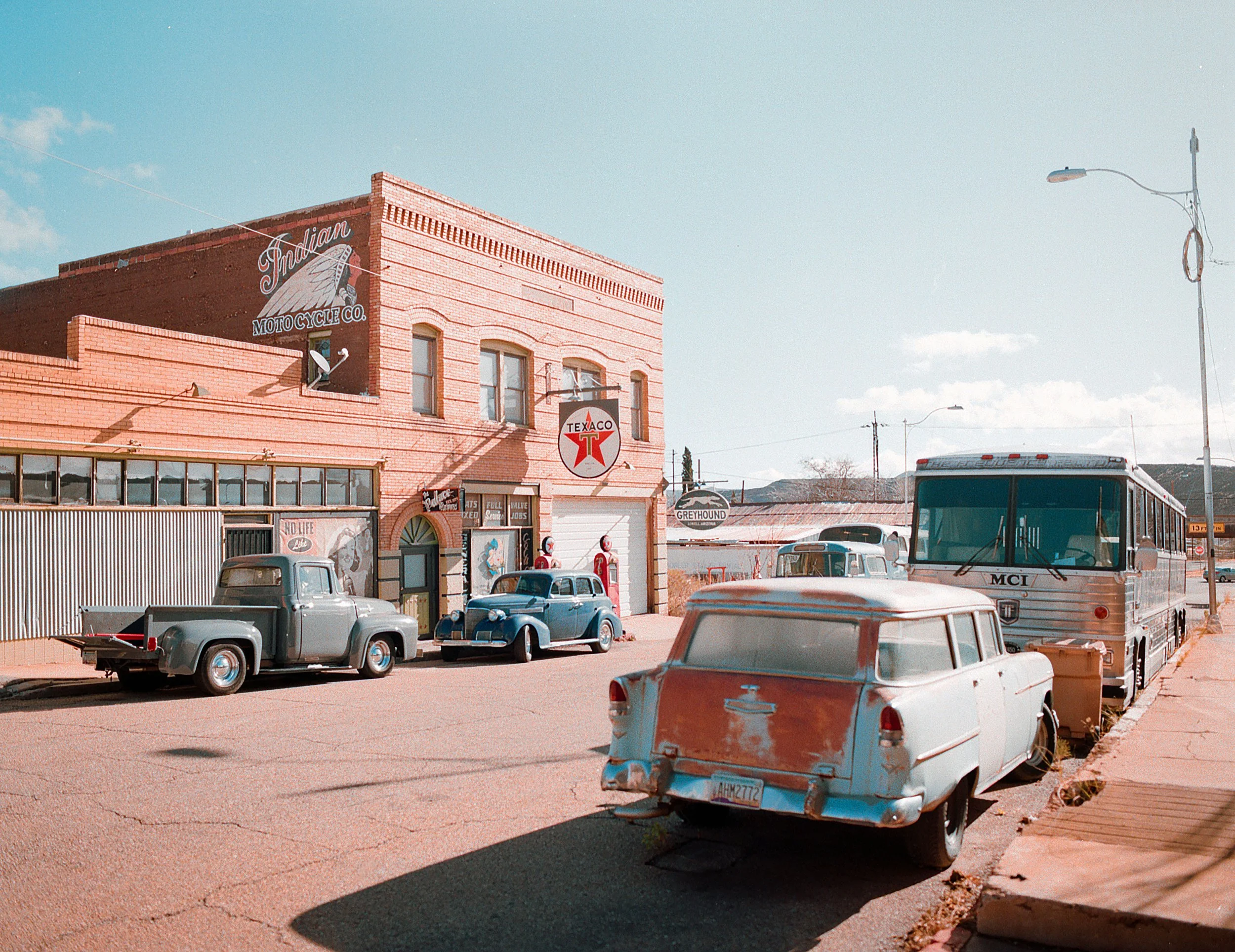 Vintage cars parked in front of an old building with signs reading 'Indian Motorcycle Co.', 'Texaco', and 'Greyhound'.