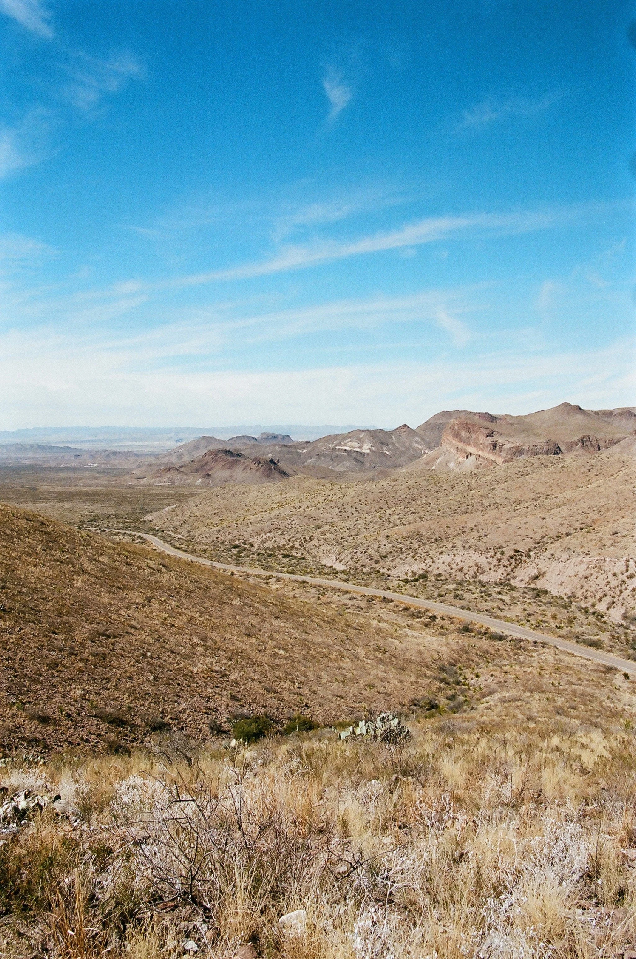 Desert landscape with rolling hills, sparse vegetation, and a winding dirt road under a blue sky with a few wispy clouds.