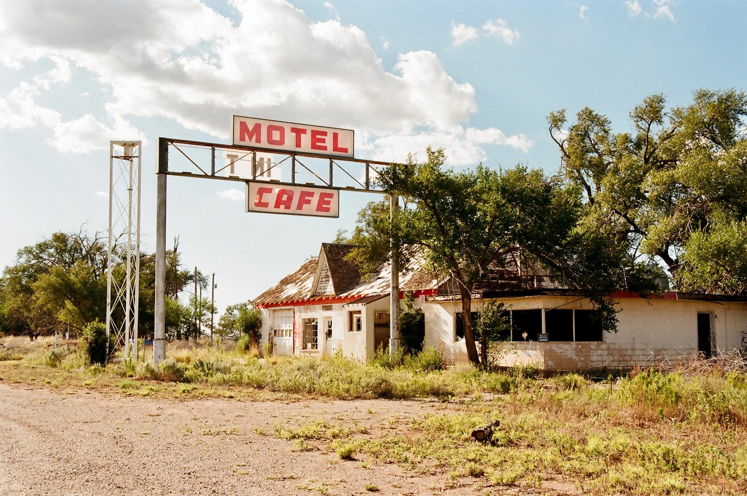 An old, abandoned building with a sign reading 'Motel Cafe' outside on a dirt road under a partly cloudy sky, surrounded by overgrown grass and trees.