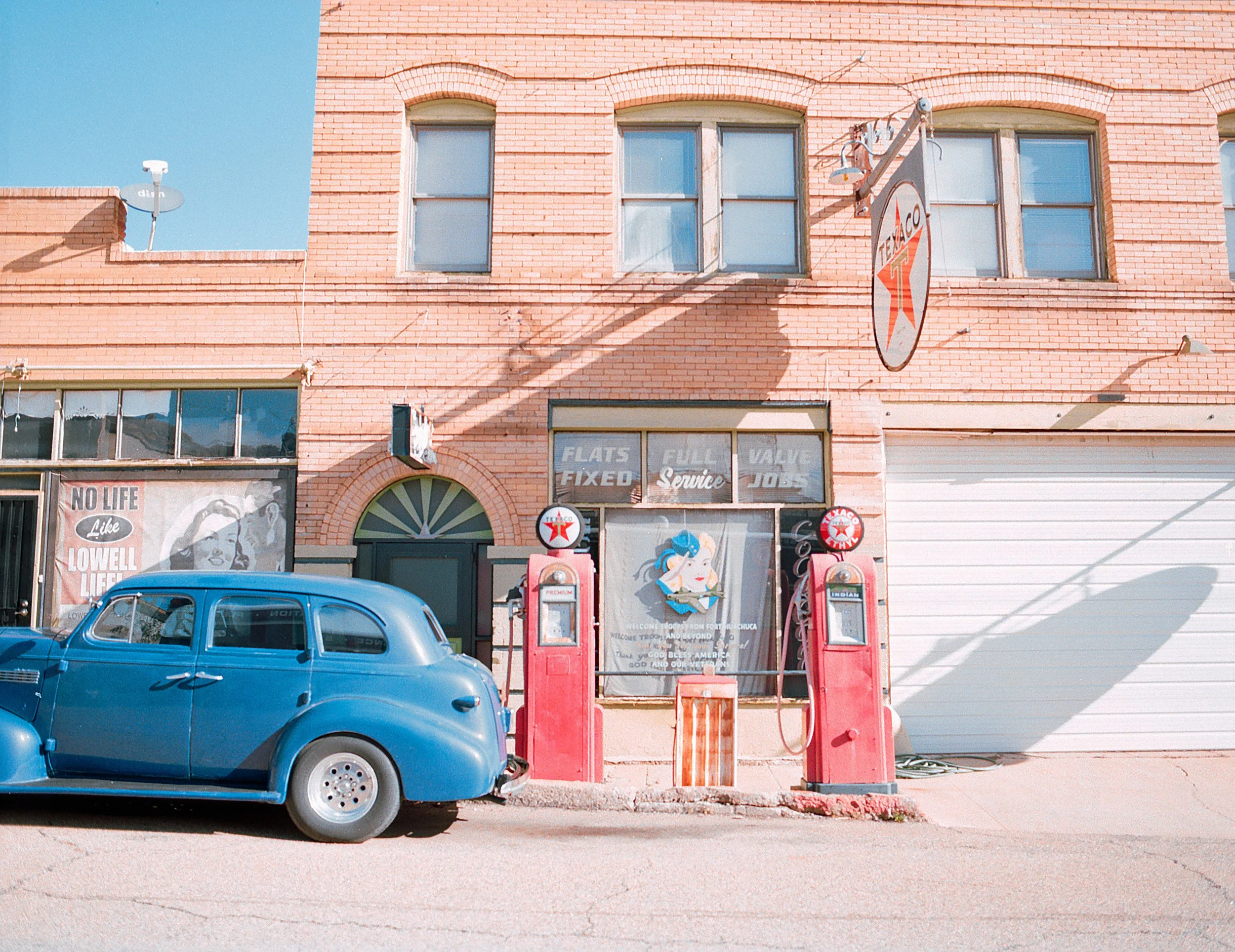 A vintage blue car parked in front of a brick building with an old gas station. The gas station has two red vintage gas pumps, and a sign for Texaco. There are windows with advertisements and a large garage door on the right. The blue sky is visible 