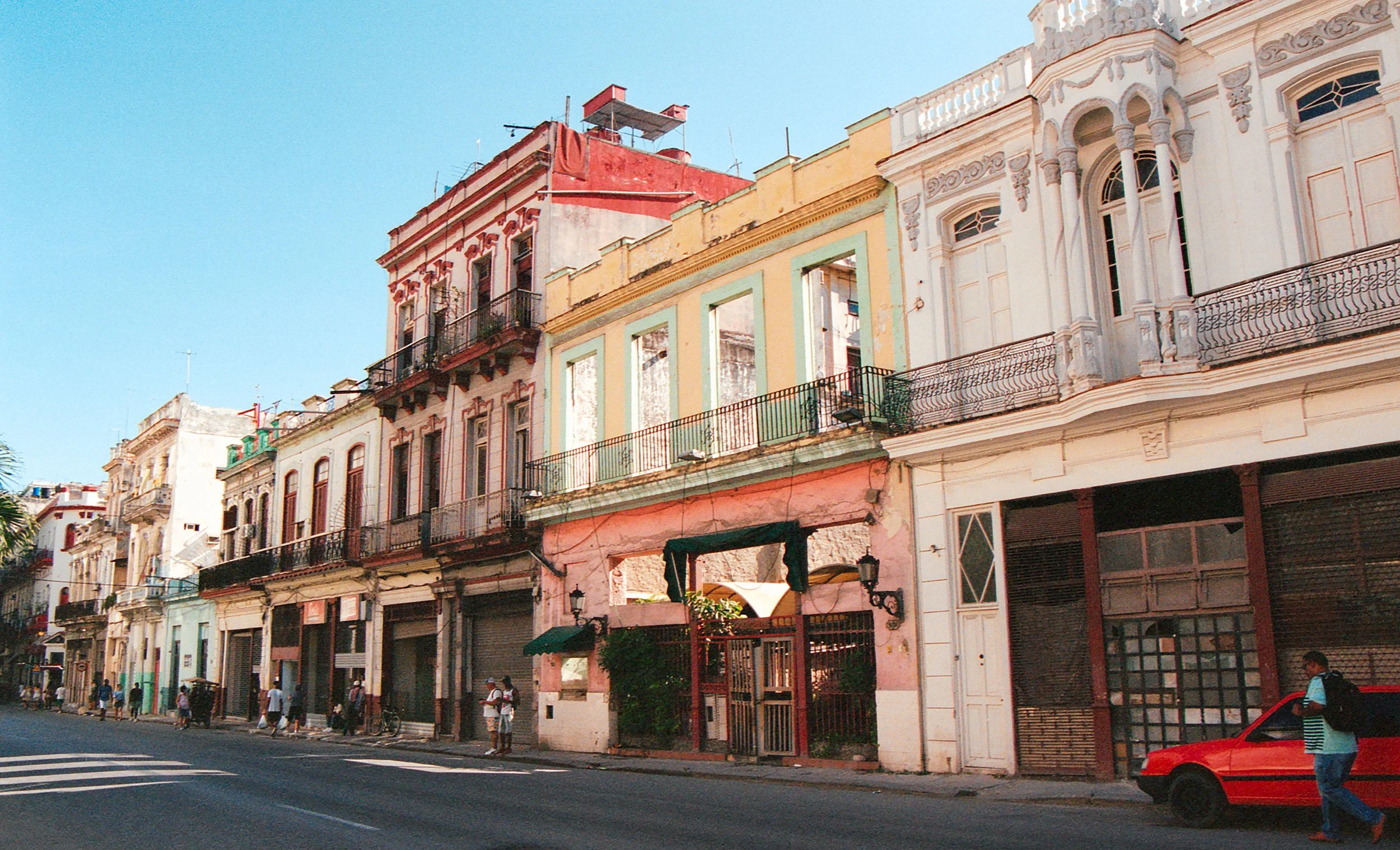 Colorful classical buildings with balconies on a city street under a clear blue sky, pedestrians walking, and a red car parked on the sidewalk.