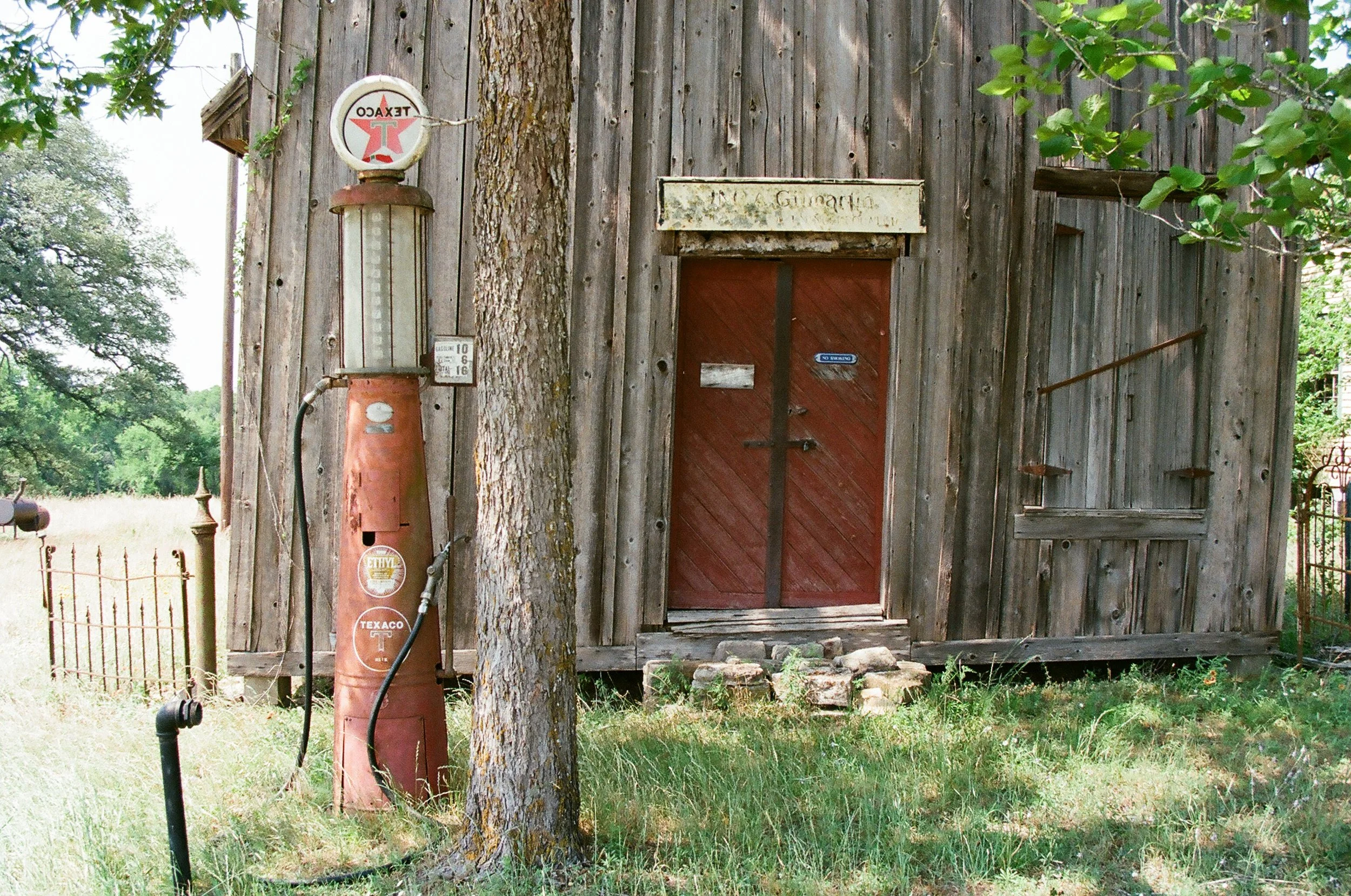 An old wooden building with a red door, a vintage gas pump, and a tree in front. The gas pump has the Texaco logo and is weathered. The building has a weathered sign above the door, and steps made of bricks lead up to it. Trees and grass surround the