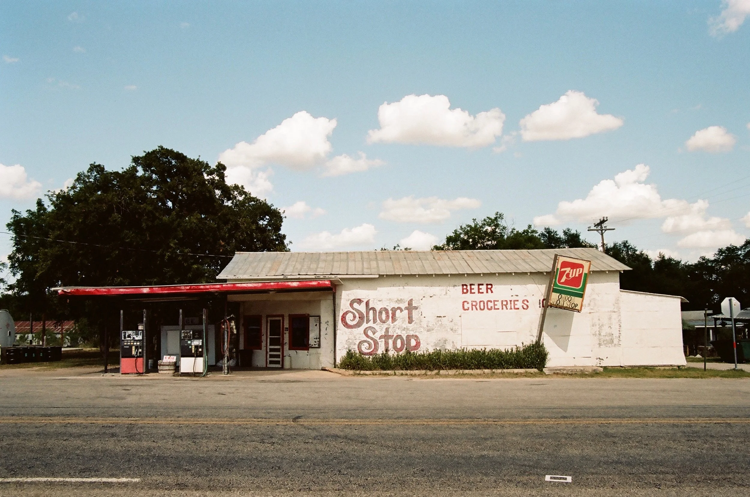 An old, small grocery store and convenience shop called Short Stop with a 7UP sign in front. The building has a white-painted brick exterior with a metal roof and red accents around the door and windows. The store's signage includes the words 'BEER',