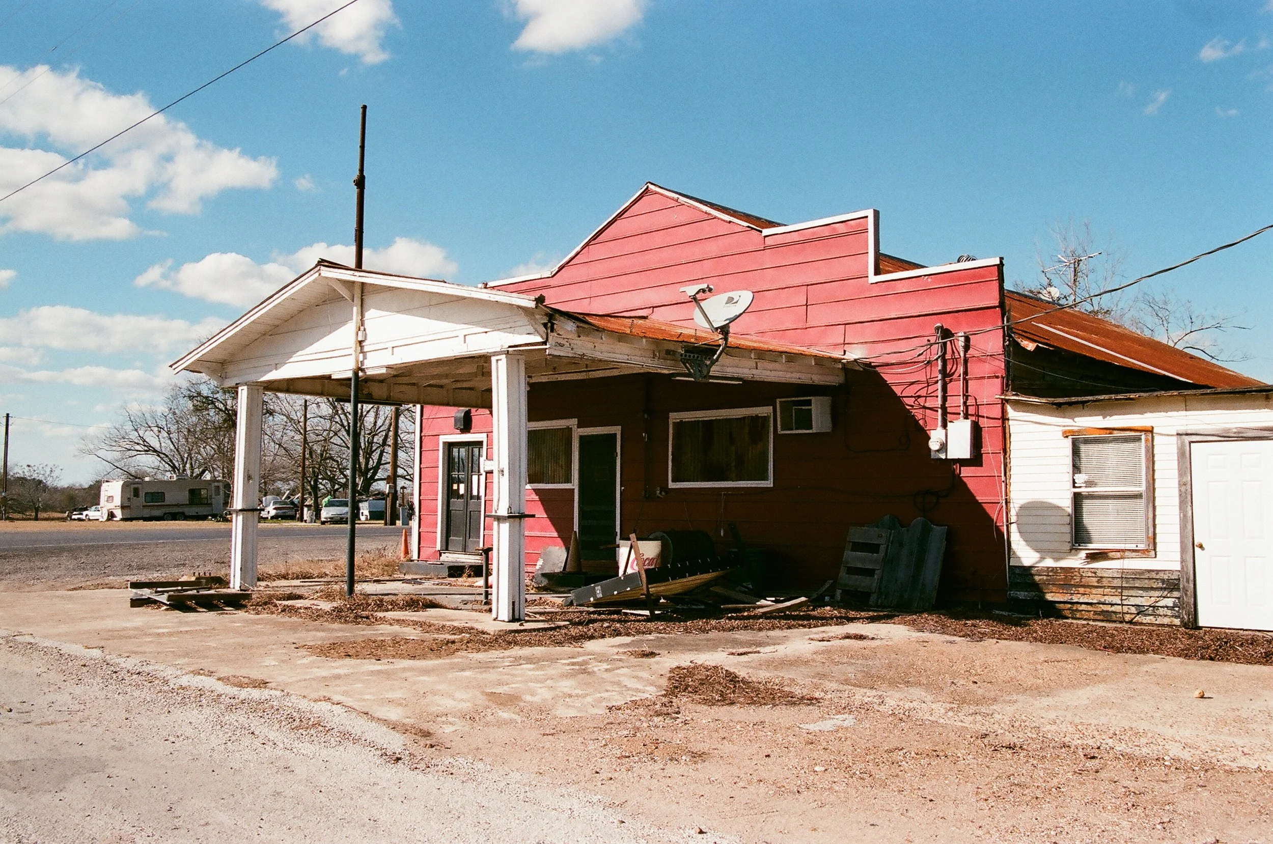 A small, old, red building with a white porch and a white attached section, situated in a rural area with a gravel ground, under a blue sky with scattered clouds; some objects and debris are on the ground near the building.