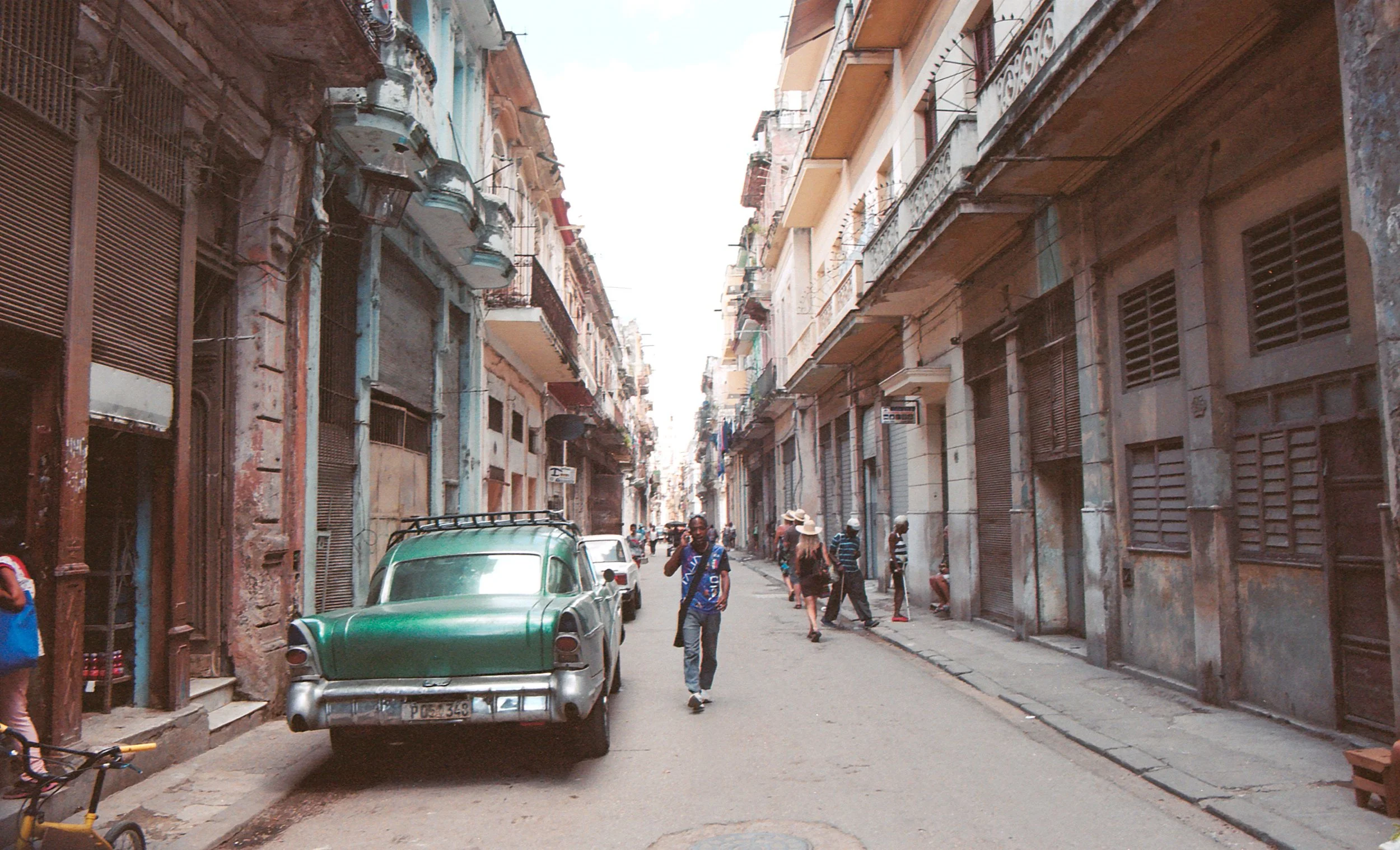 A narrow street in an old neighborhood with colorful, weathered buildings, several pedestrians walking, and a vintage green car parked on the side.