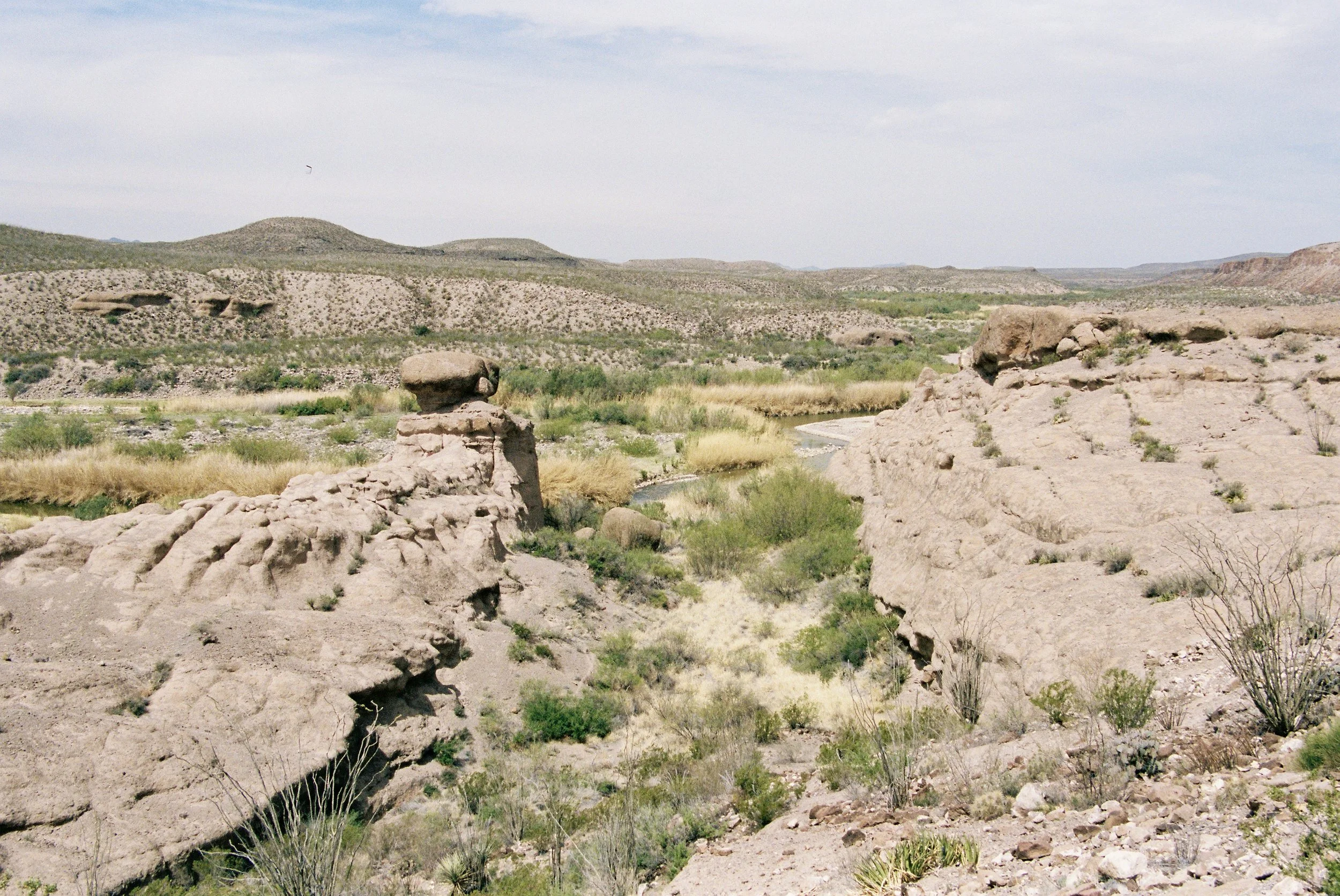 Arid desert landscape with large rock formations, sparse green vegetation, a small river or creek in the valley, and hills in the background under a partly cloudy sky.