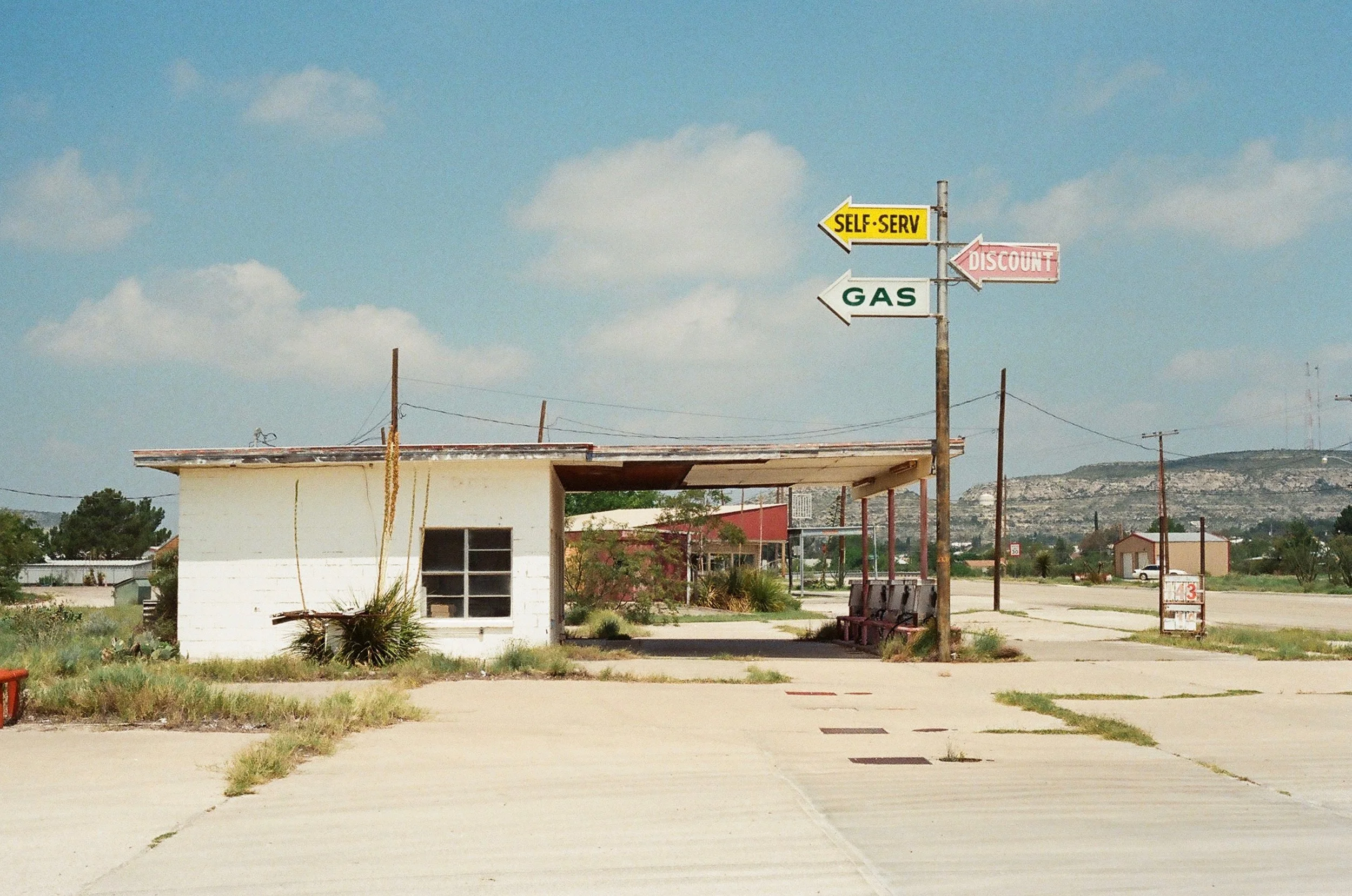 An old, abandoned gas station with three colorful signs pointing in different directions: yellow for self-serve, green for gas, and pink for discount. The station has a small white building with a window, overgrown plants, and a bench. The background
