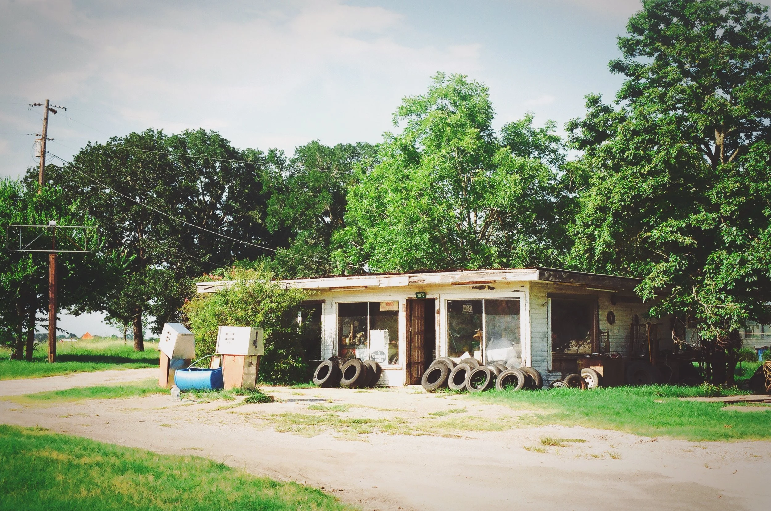 An old, small, white building in a rural setting with large green trees behind it. There are old tires in front of the building and two mailboxes near the path. The sky is partly cloudy.