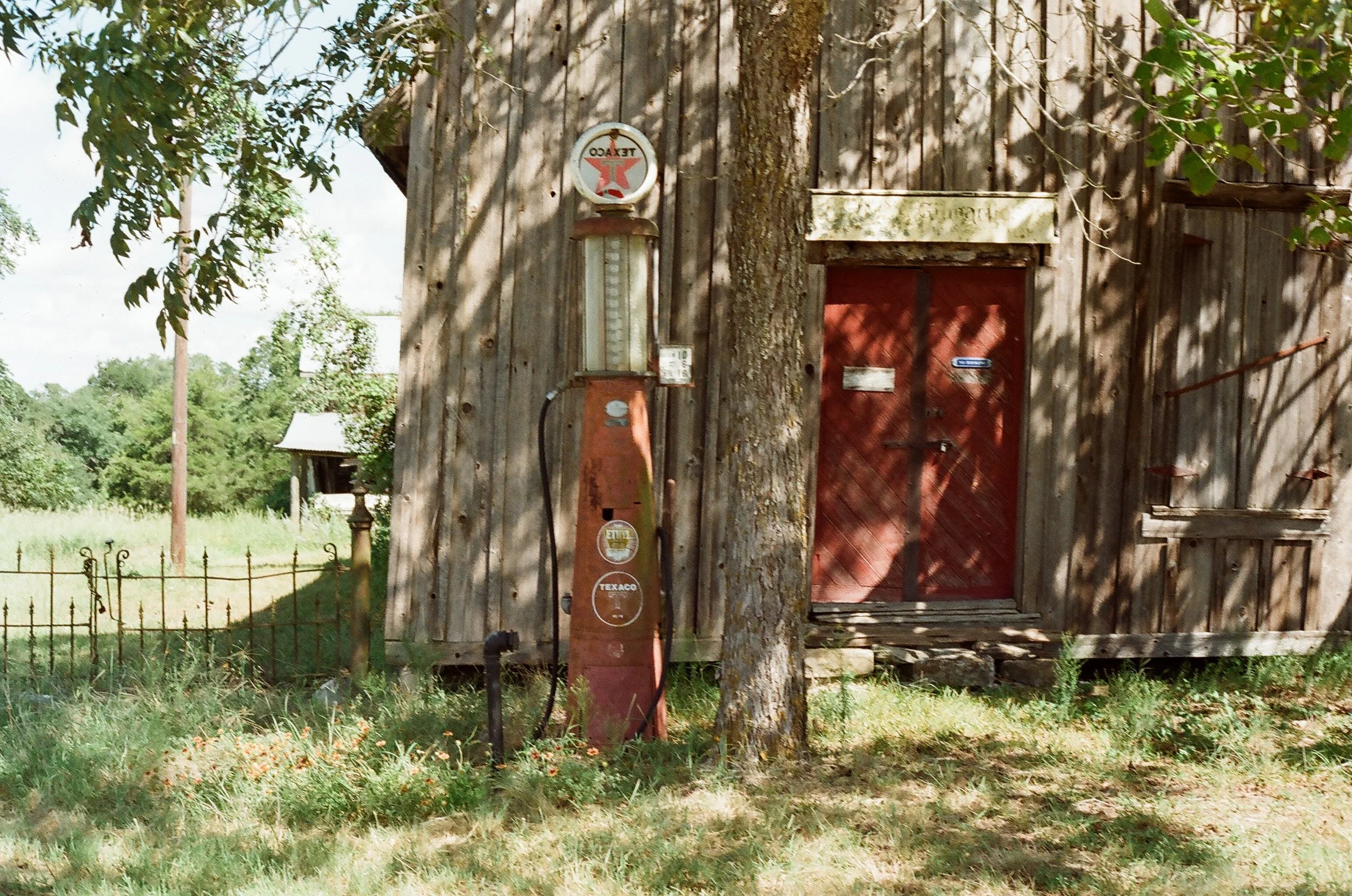 Old wooden building with a red door and vintage Texaco gas pump beside a tree in a grassy yard.
