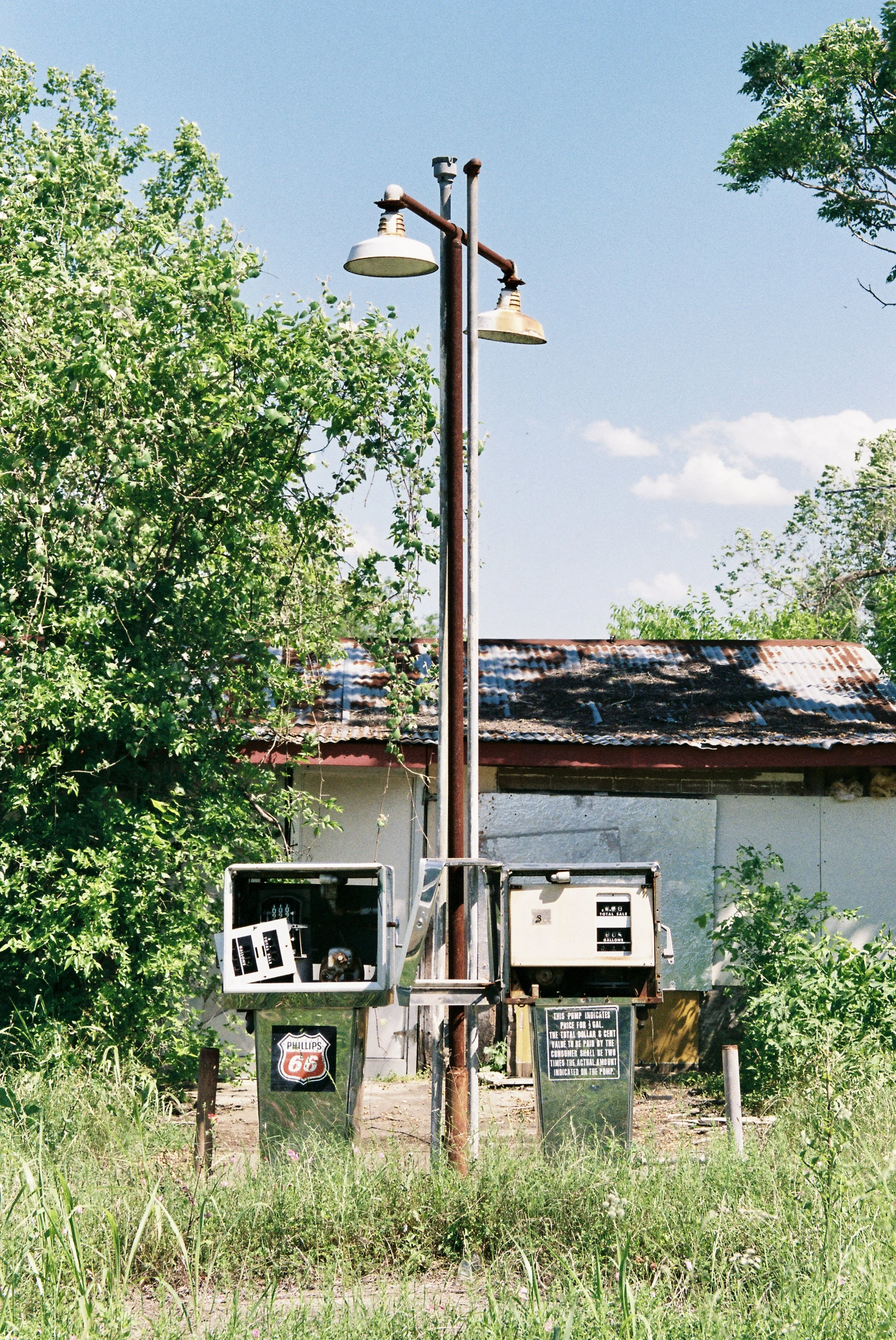 Vintage gas station meters and lamps attached to a rusted pole, with overgrown grass and an old building in the background under a blue sky.