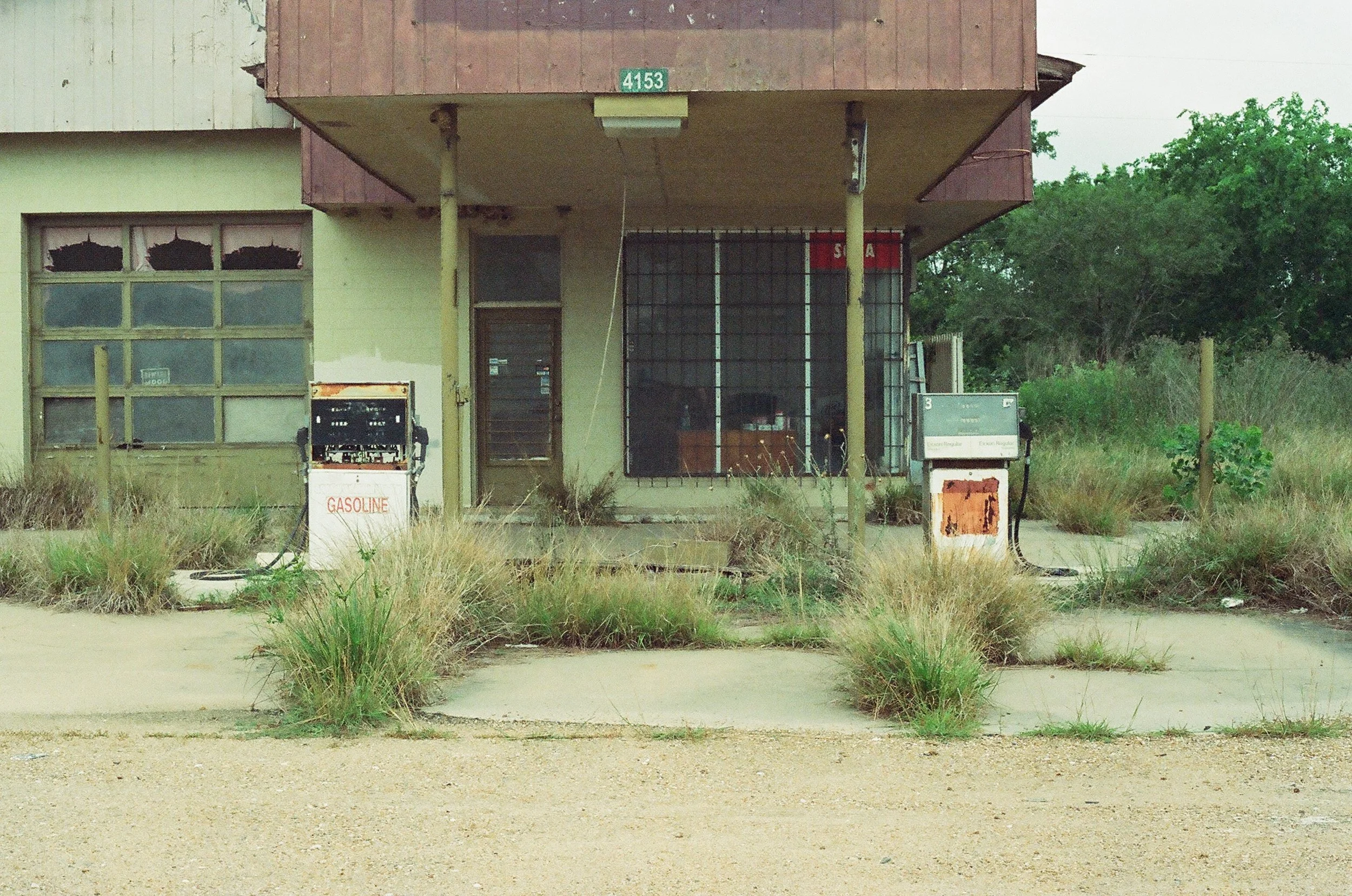 Abandoned gas station with two gas pumps, overgrown grass, and an old building with boarded-up windows and a metal grate covering the entrance.