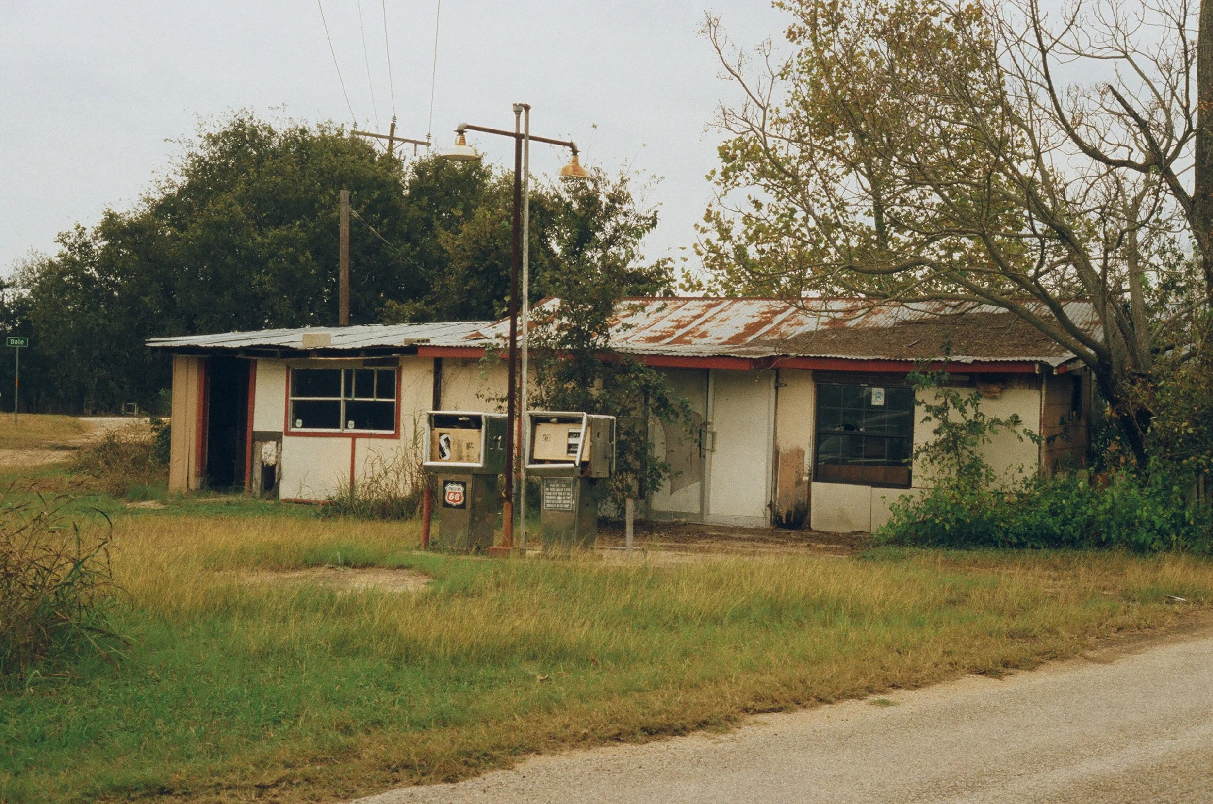 An old, rundown building with a rusted metal roof, surrounded by overgrown grass and trees, with two outdoor mailboxes in front and a street sign on the corner.