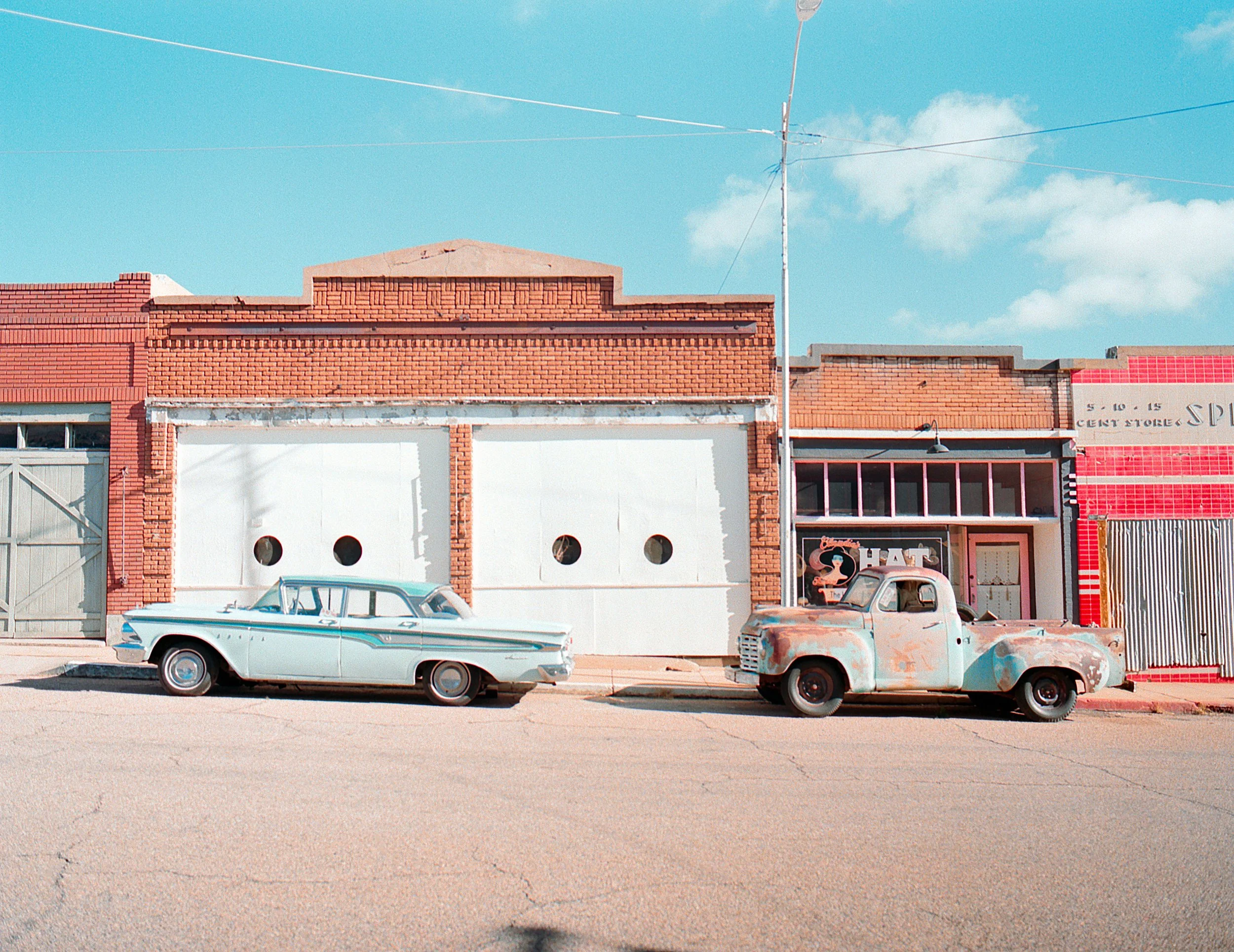 Old, weathered cars parked in front of brick buildings on a sunny day with a clear blue sky.