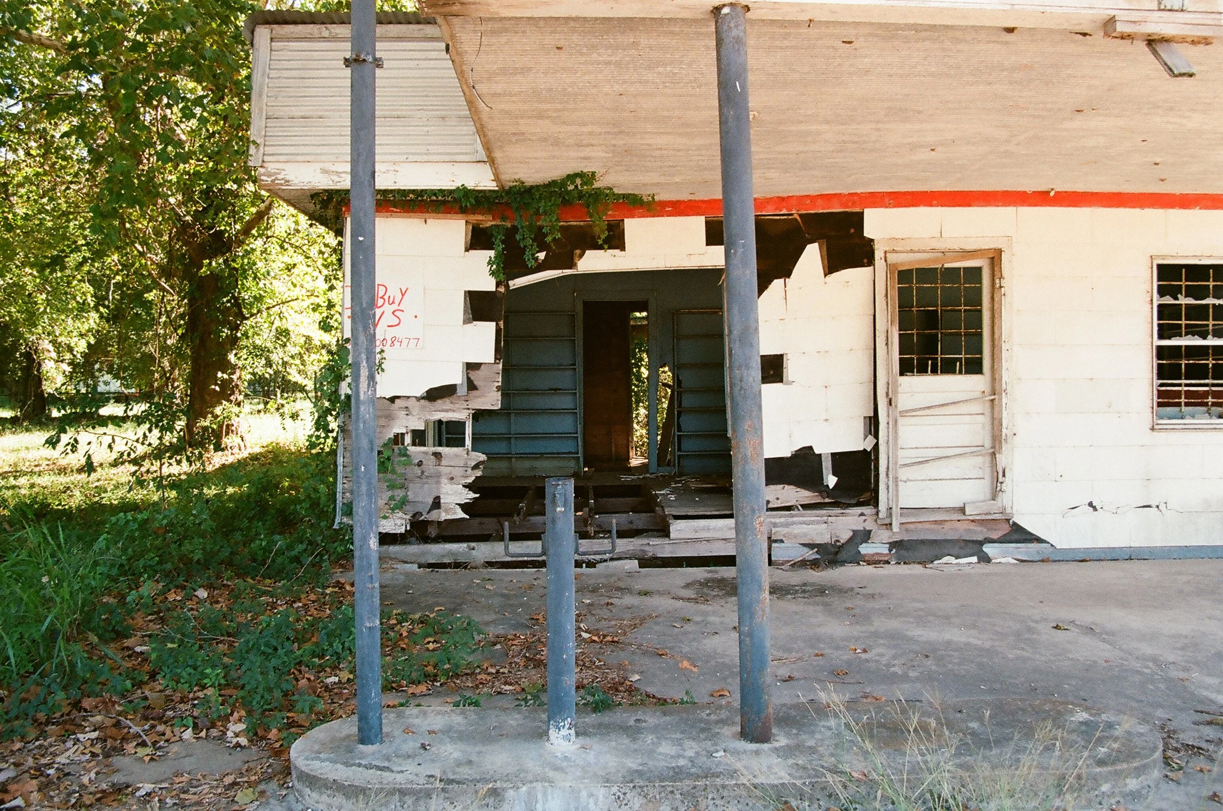 An abandoned building with a partially broken porch and dilapidated door and windows, surrounded by overgrown vegetation and fallen leaves.