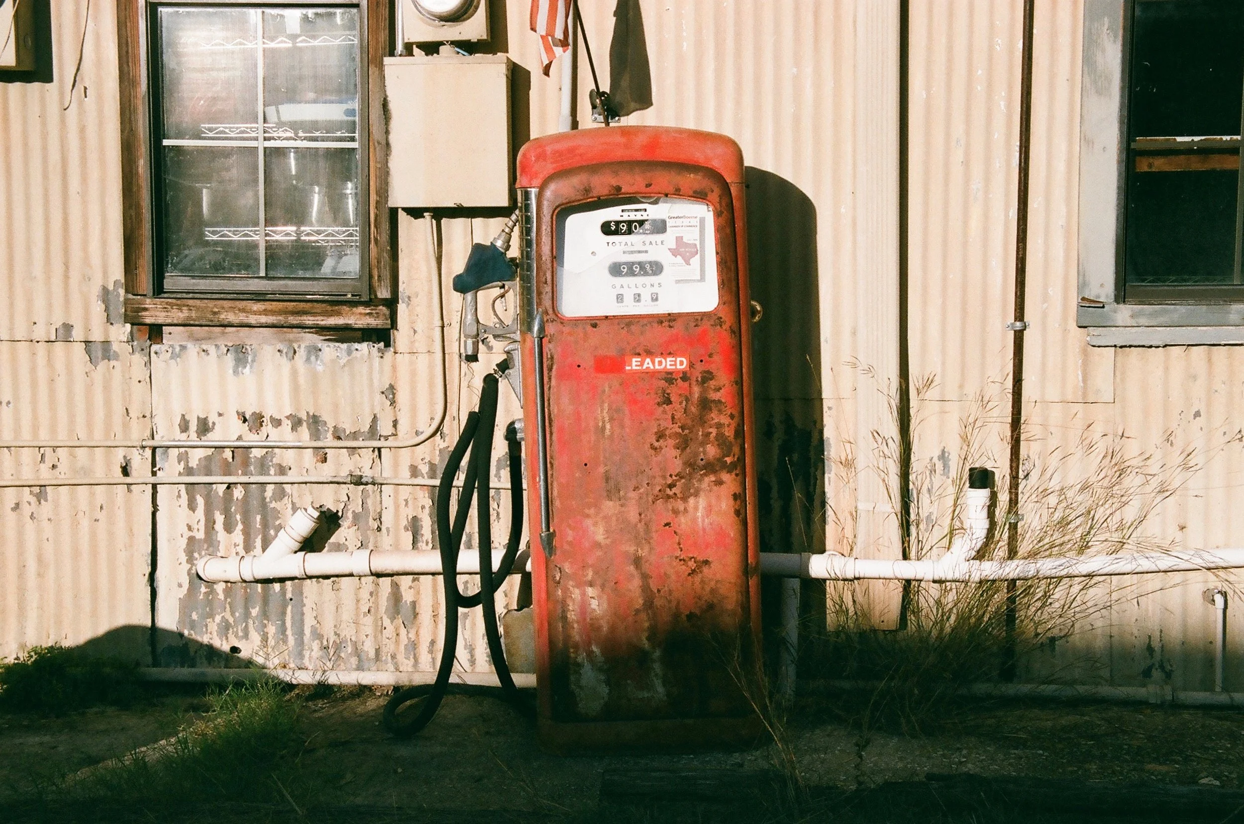An old, red gas pump in front of a weathered, beige corrugated metal building with two small windows. The gas pump shows signs of rust and wear, surrounded by grass and dirt.