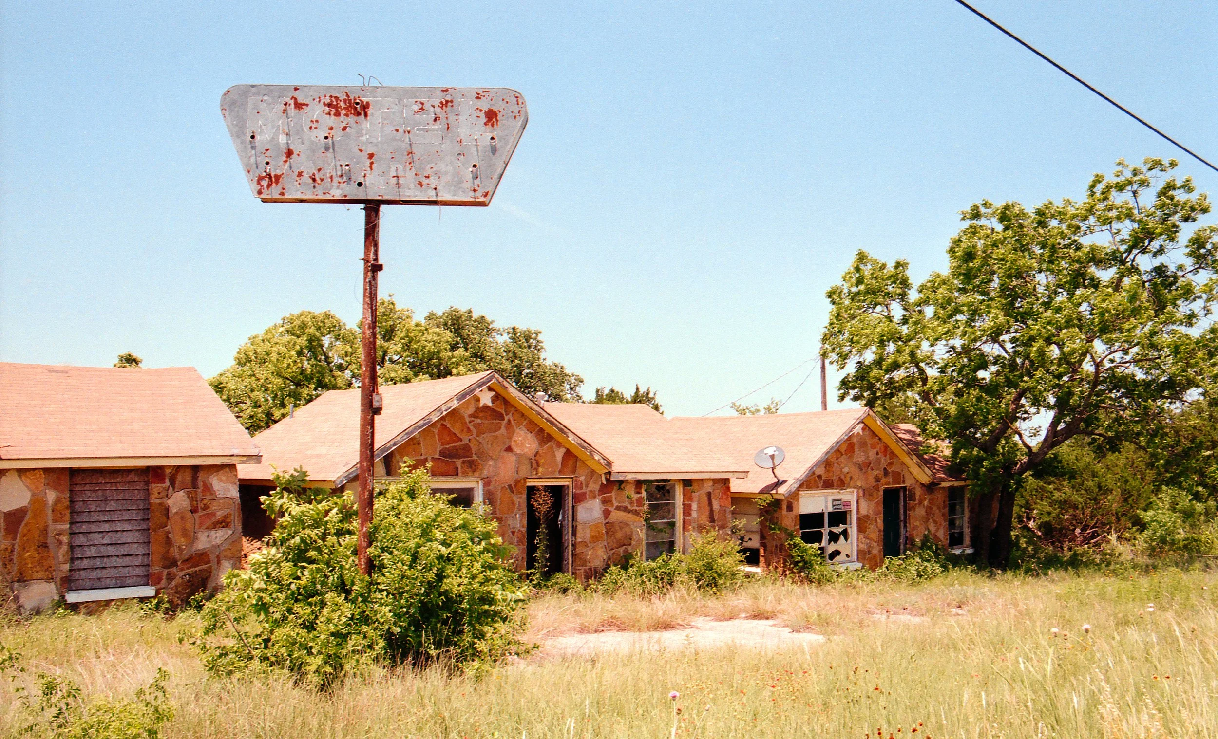 Abandoned stone house with broken windows, overgrown grass and trees, rusty sign, and a satellite dish in a rural setting under clear blue sky.