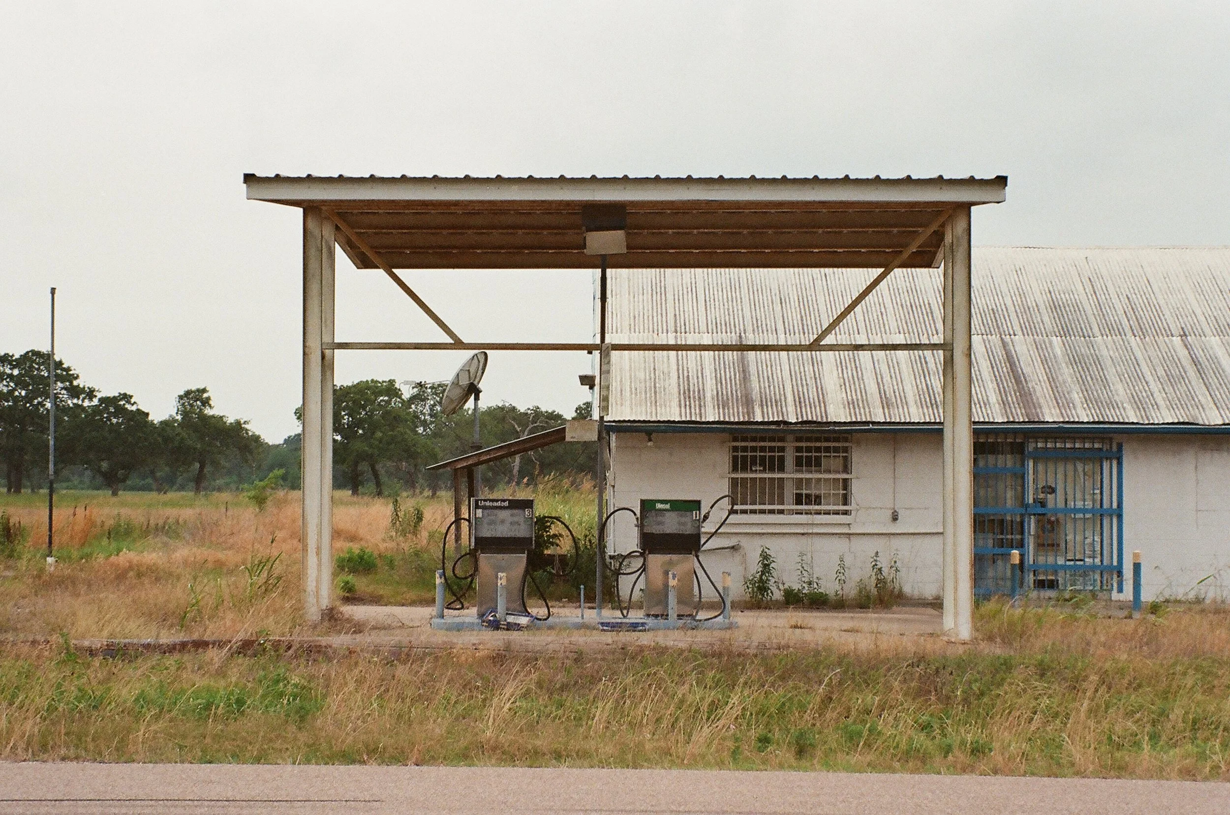 A small roadside fuel station with two fuel pumps labeled 'Unleaded' and 'Diesel', attached to a simple wooden structure with a metal roof, surrounded by grass and a rural landscape.