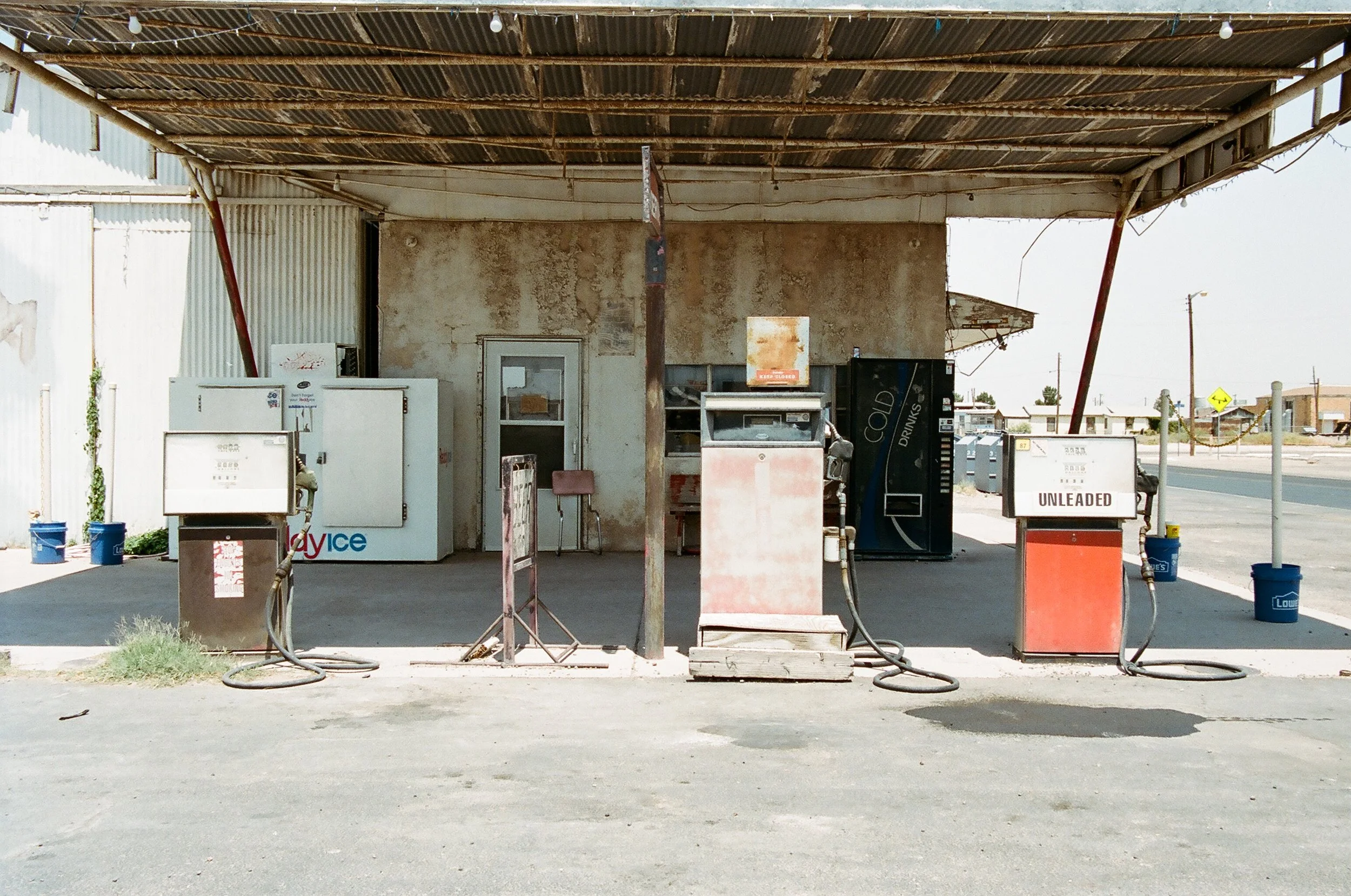 Old gas station with two vintage fuel pumps labeled 'Unleaded' and 'Vice', situated beneath a rusted metal canopy. The station has a weathered, peeling wall with a small glass door leading inside. There is a black vending machine labeled 'Cold Drinks