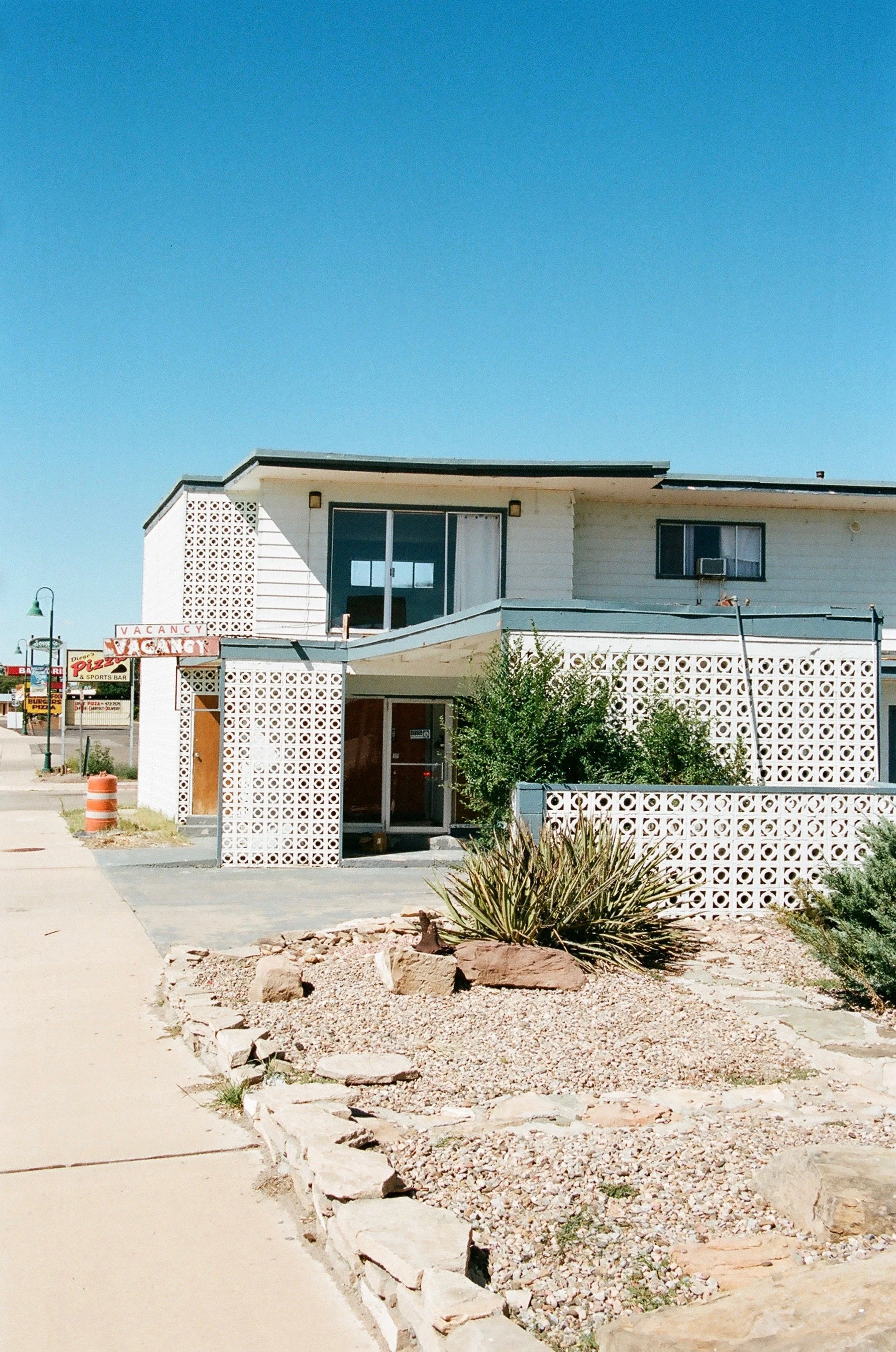 A two-story building with white exterior walls and decorative white brickwork. There are large windows on the upper floor and a glass door on the ground floor, surrounded by desert landscaping with rocks and sparse plants. A sign indicates vacancy fo