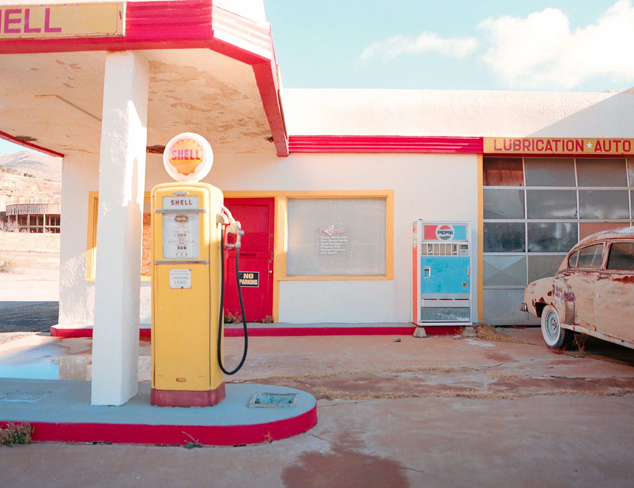 Old vintage gas station with yellow Shell pump, red and white building, and a rusty car on the right. The building has signs reading 'Lubrication Auto' and a vending machine.