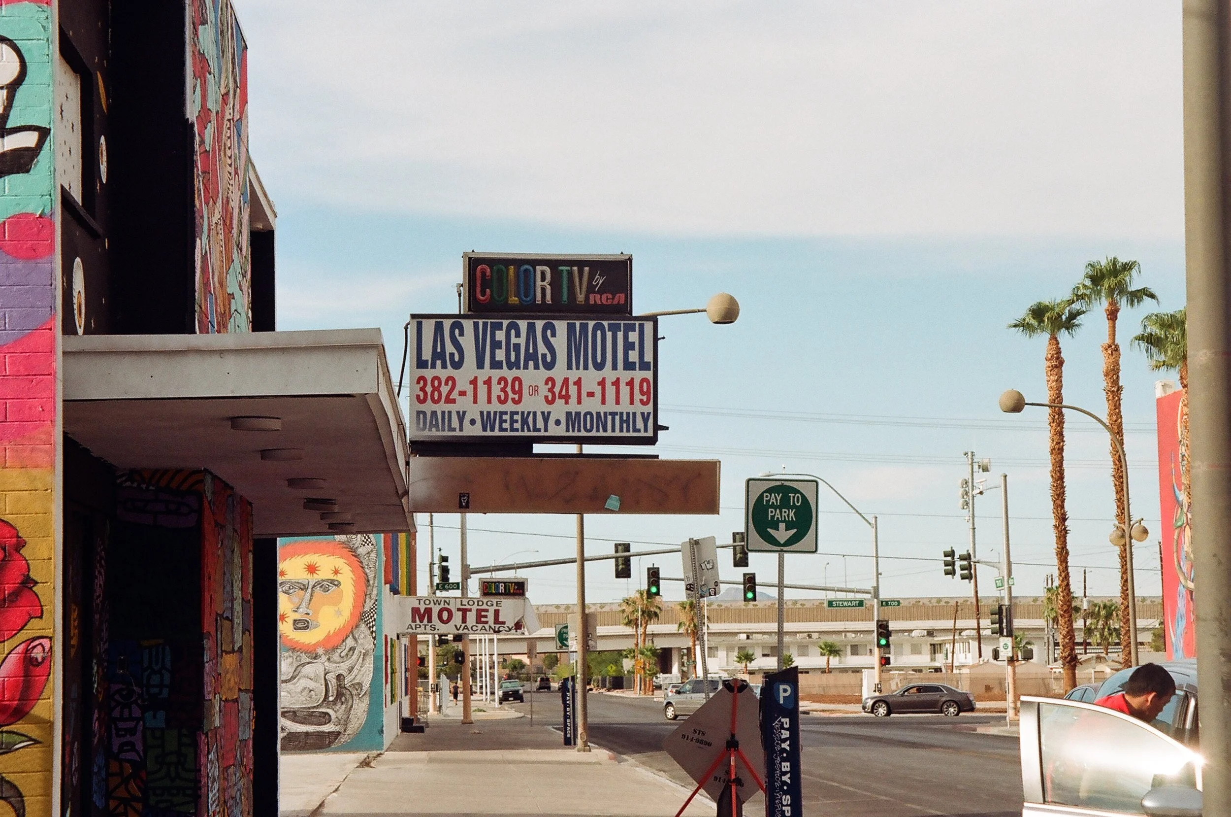 Street view of Las Vegas with signs for a motel, palm trees, colorful murals, and a man near a car.