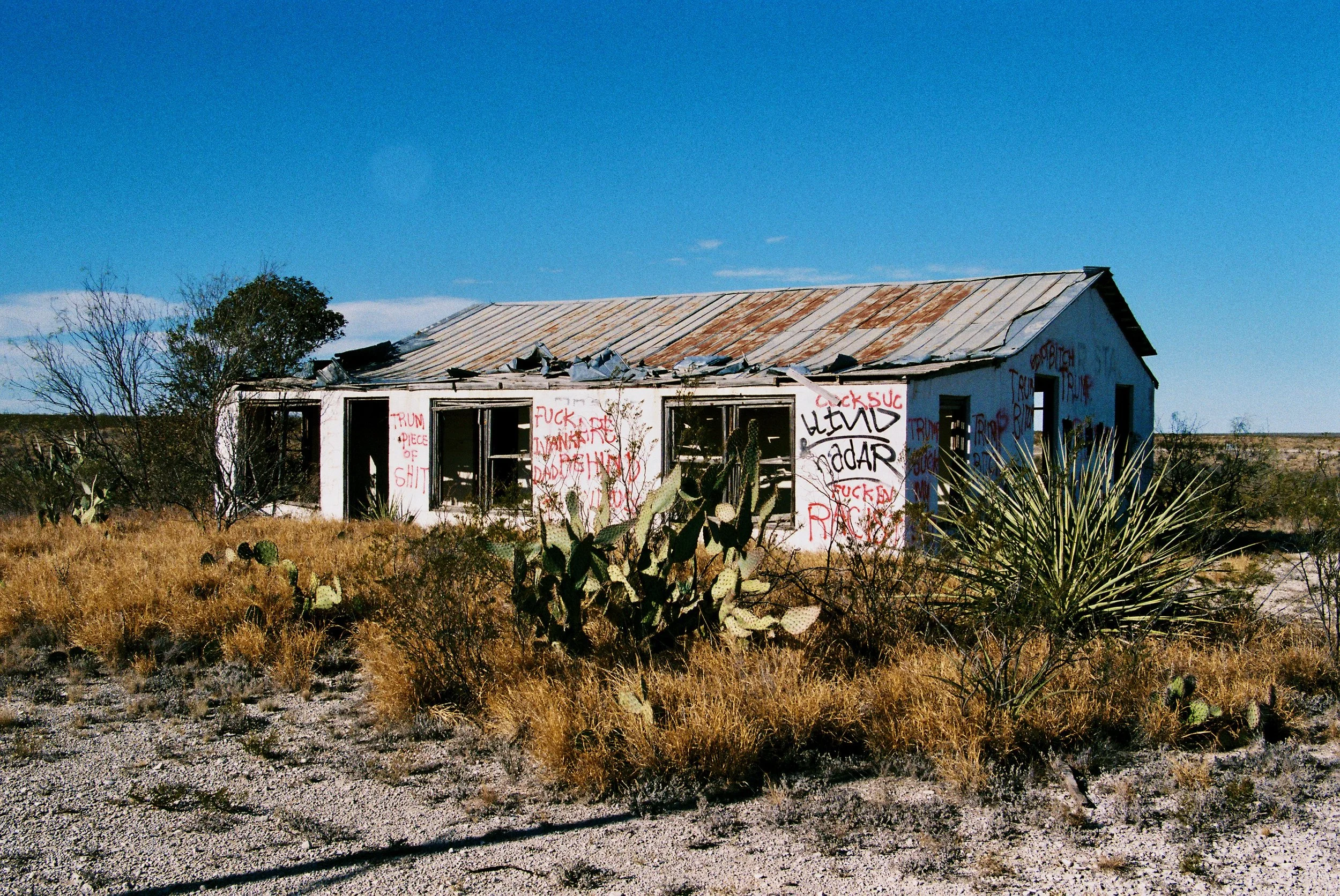 Abandoned house in a desert landscape with graffiti written in red spray paint on its walls, surrounded by dry grass, cacti, and sparse shrubbery under a clear blue sky.