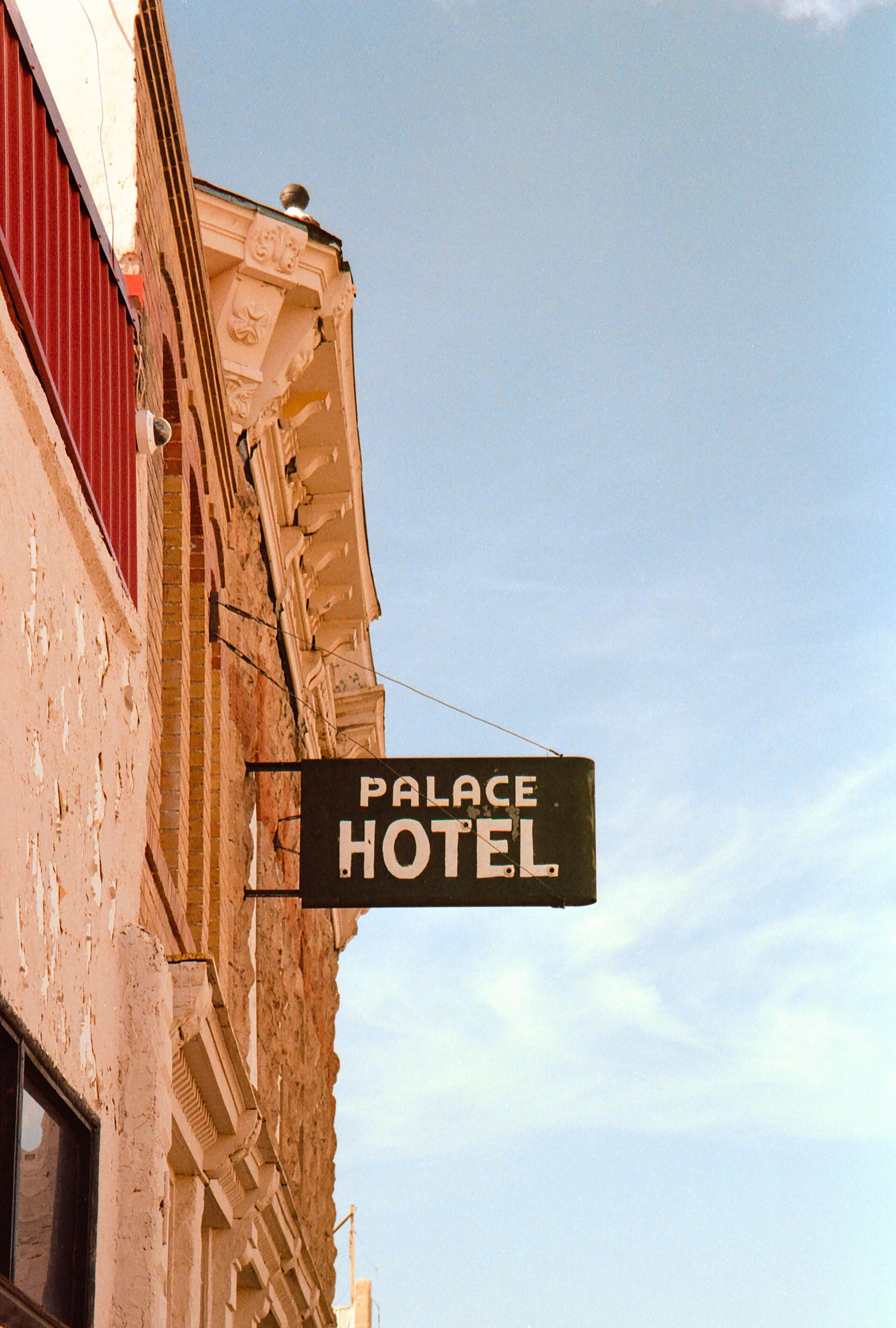 Black sign with white lettering reading "Palace Hotel" hanging outside an old building with a brick and stucco exterior and ornate architectural details, against a clear blue sky.