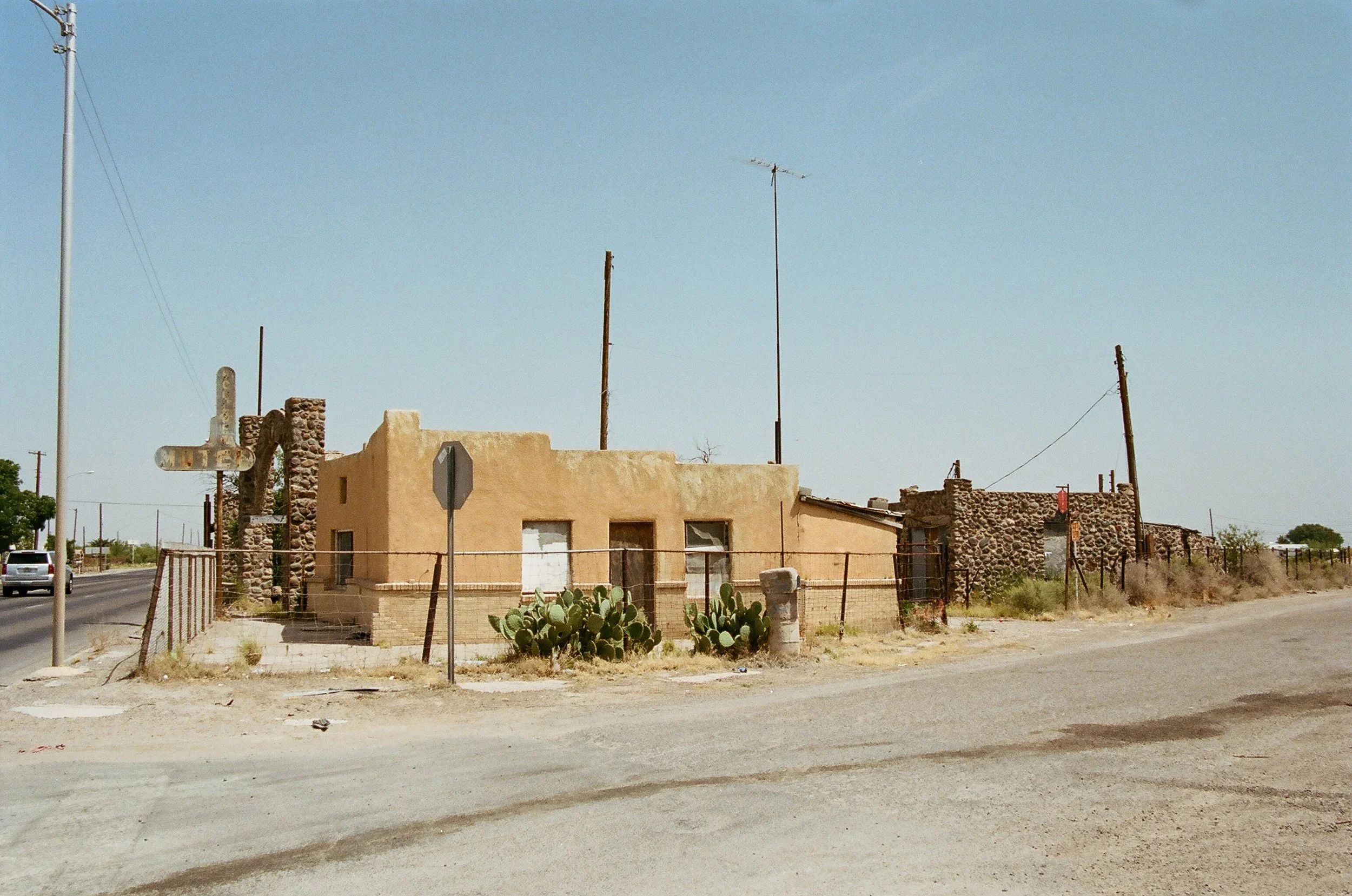 A weathered, adobe-style house with cactus in front, surrounded by a wire fence, set in a dry, dusty landscape with power lines and a stop sign nearby, under a clear blue sky.