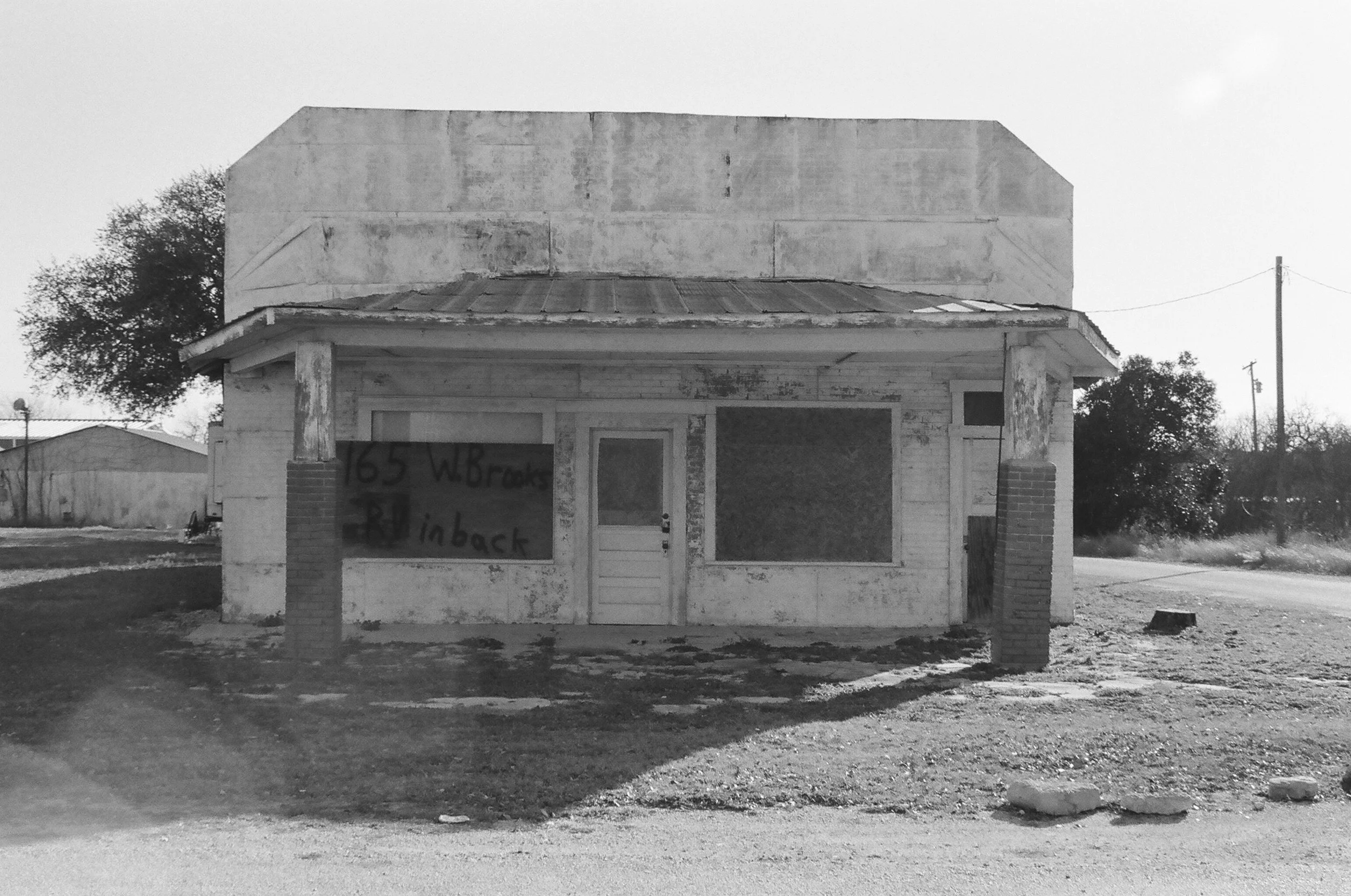 An abandoned, small, two-story building with peeling paint, boarded-up windows, and a weathered front porch, situated on a dirt lot with a sign on the window reading '165 W. Brooks' and 'Rinback'.