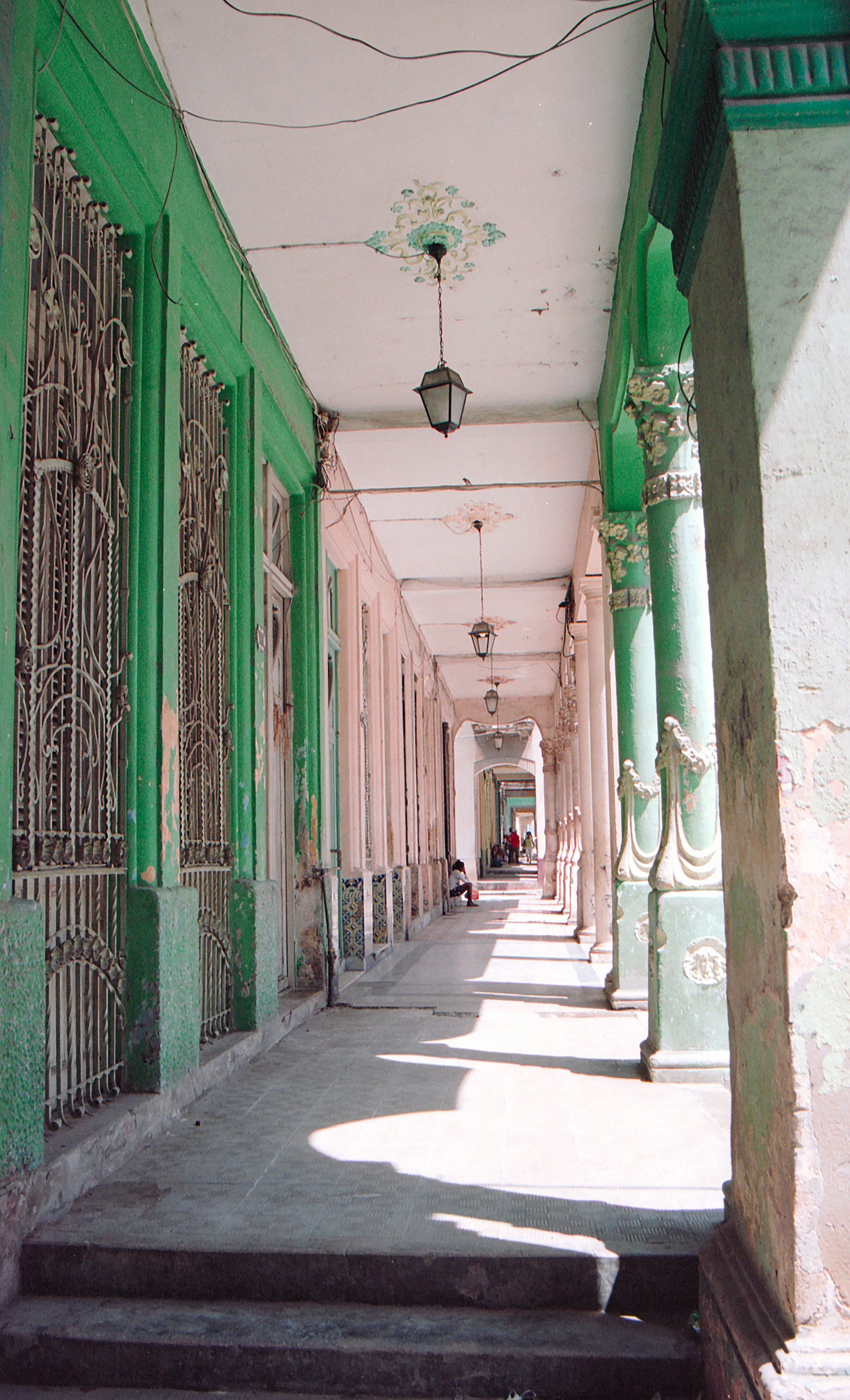Long covered outdoor walkway with green columns, decorative iron gates, and hanging lanterns, showing signs of aging and peeling paint, with a few people sitting and walking in the distance.