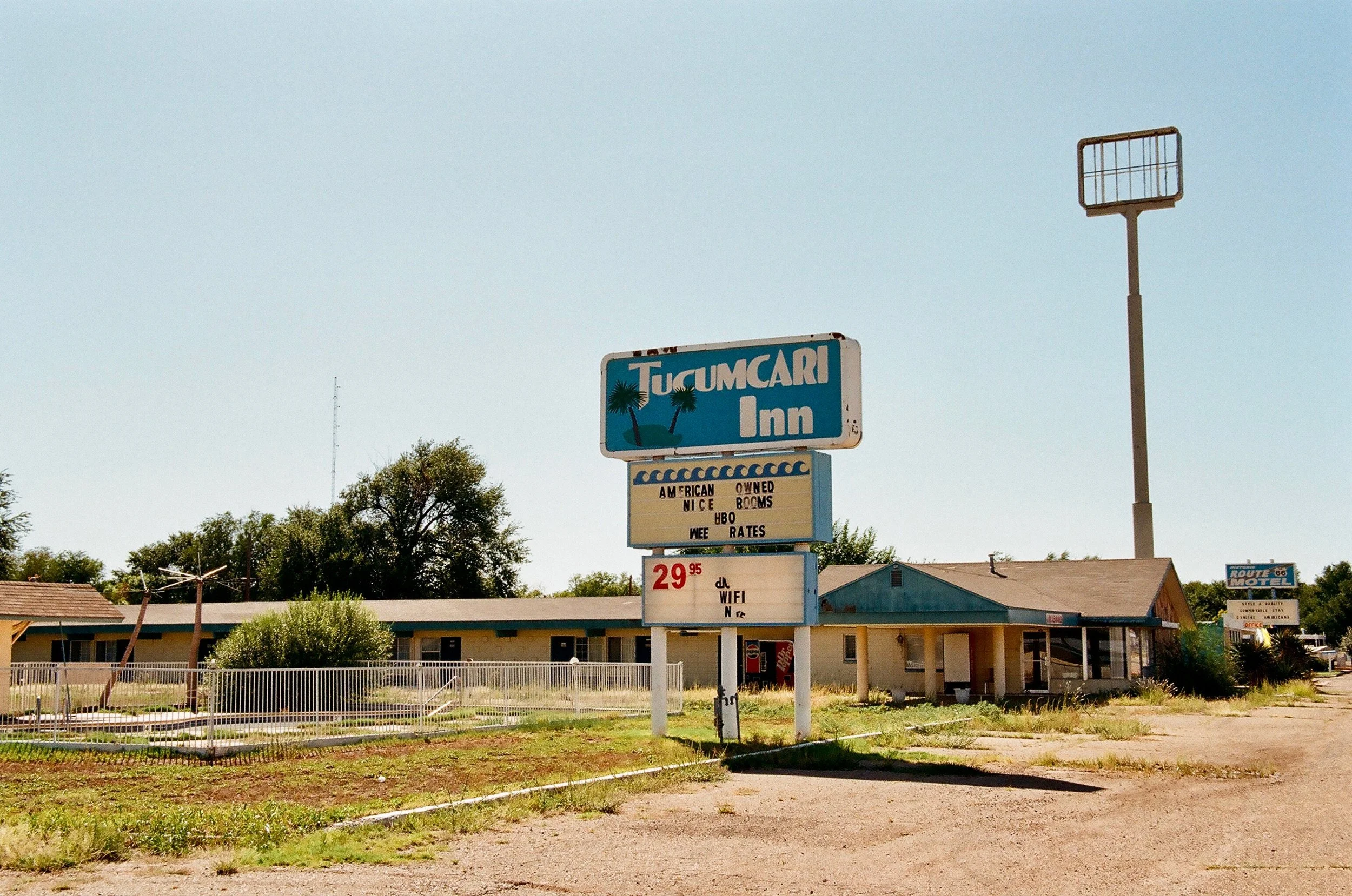 A weathered motel sign outdoors with a building in the background, advertising Tucumcari Inn with amenities and prices, in a desert-like landscape under a clear sky.