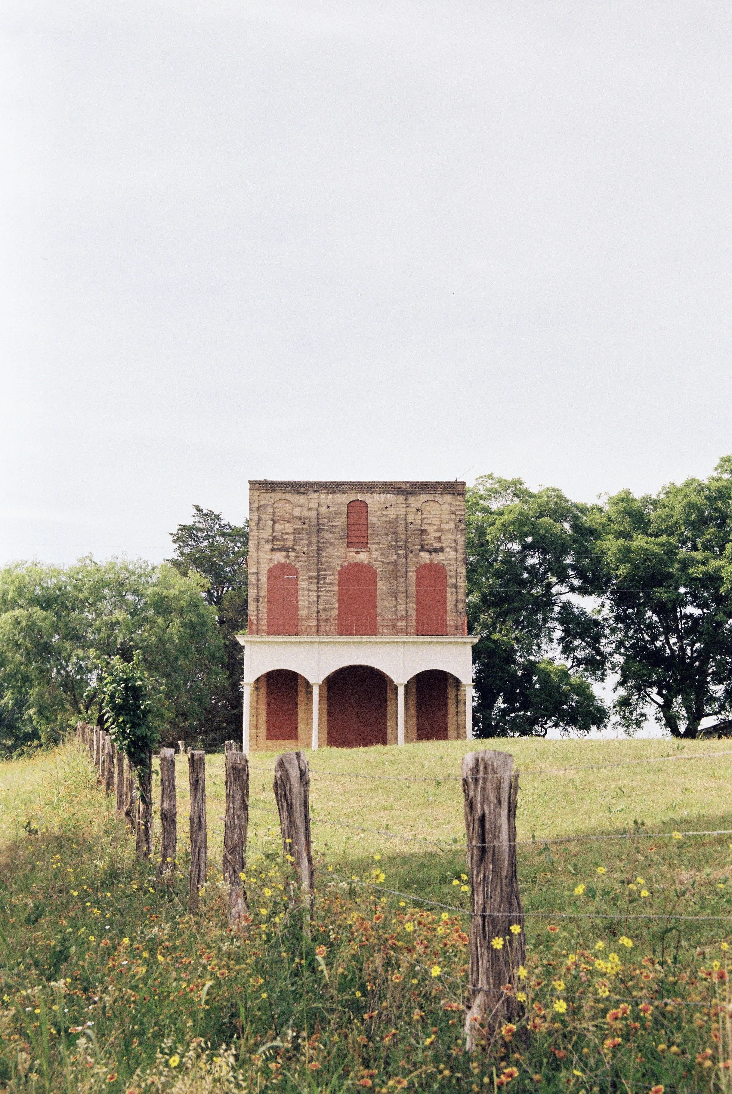 A historic brick building with an unfinished upper story, featuring three arched openings on the ground floor and boarded-up windows on the upper floor, on a grassy hill with a wooden fence and wildflowers, surrounded by trees.