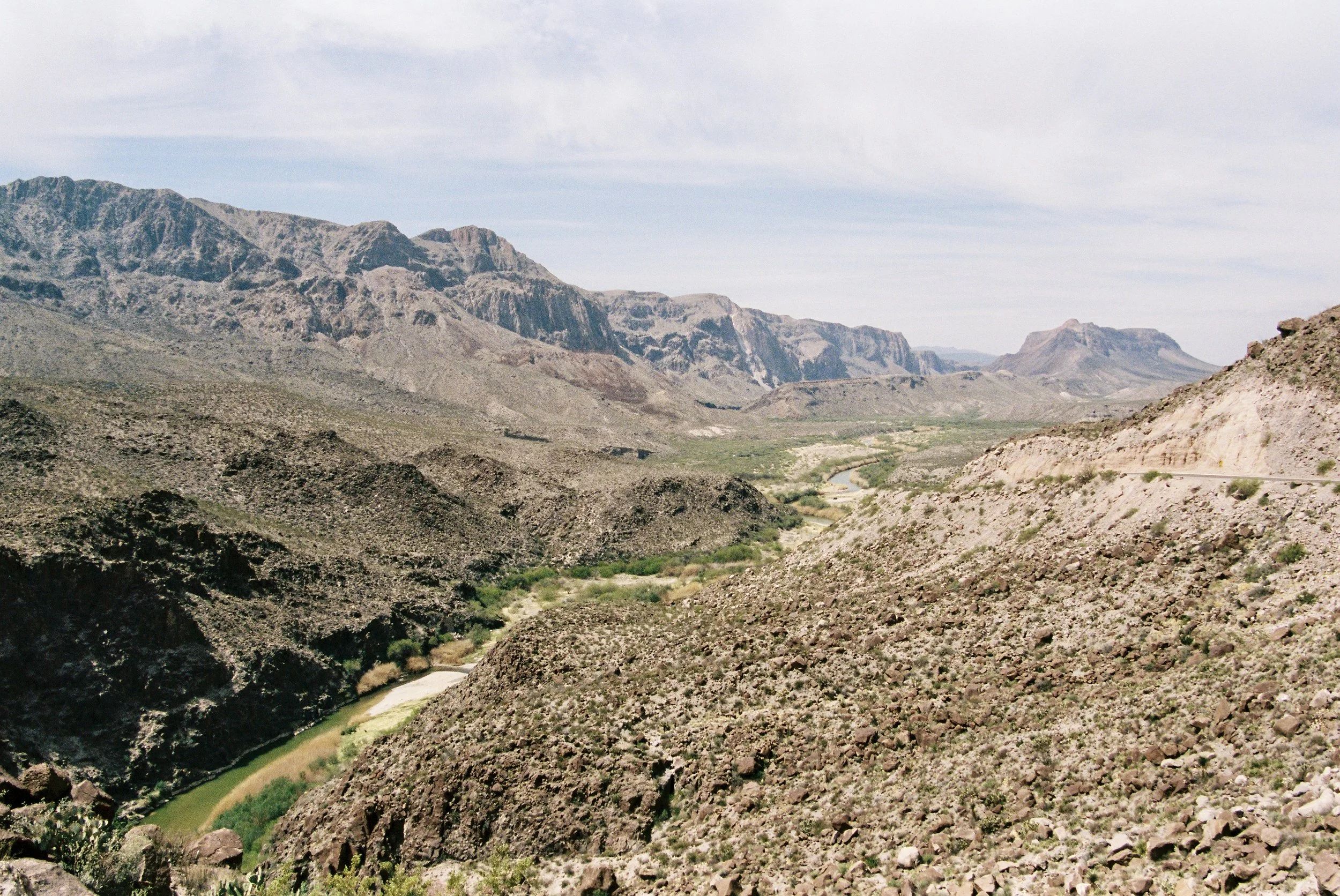 A vast desert landscape with rocky hills and mountains in the distance, featuring a meandering green river flowing through the arid terrain under a partly cloudy sky.