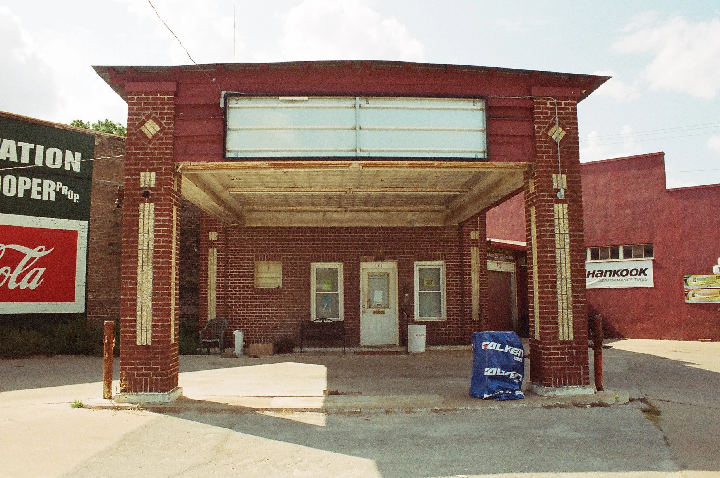 A small, old brick building with an empty outdoor canopy, a white front door, and two small windows on either side. There are signs and advertisements on neighboring walls, including Coca-Cola and Hankook Tires.