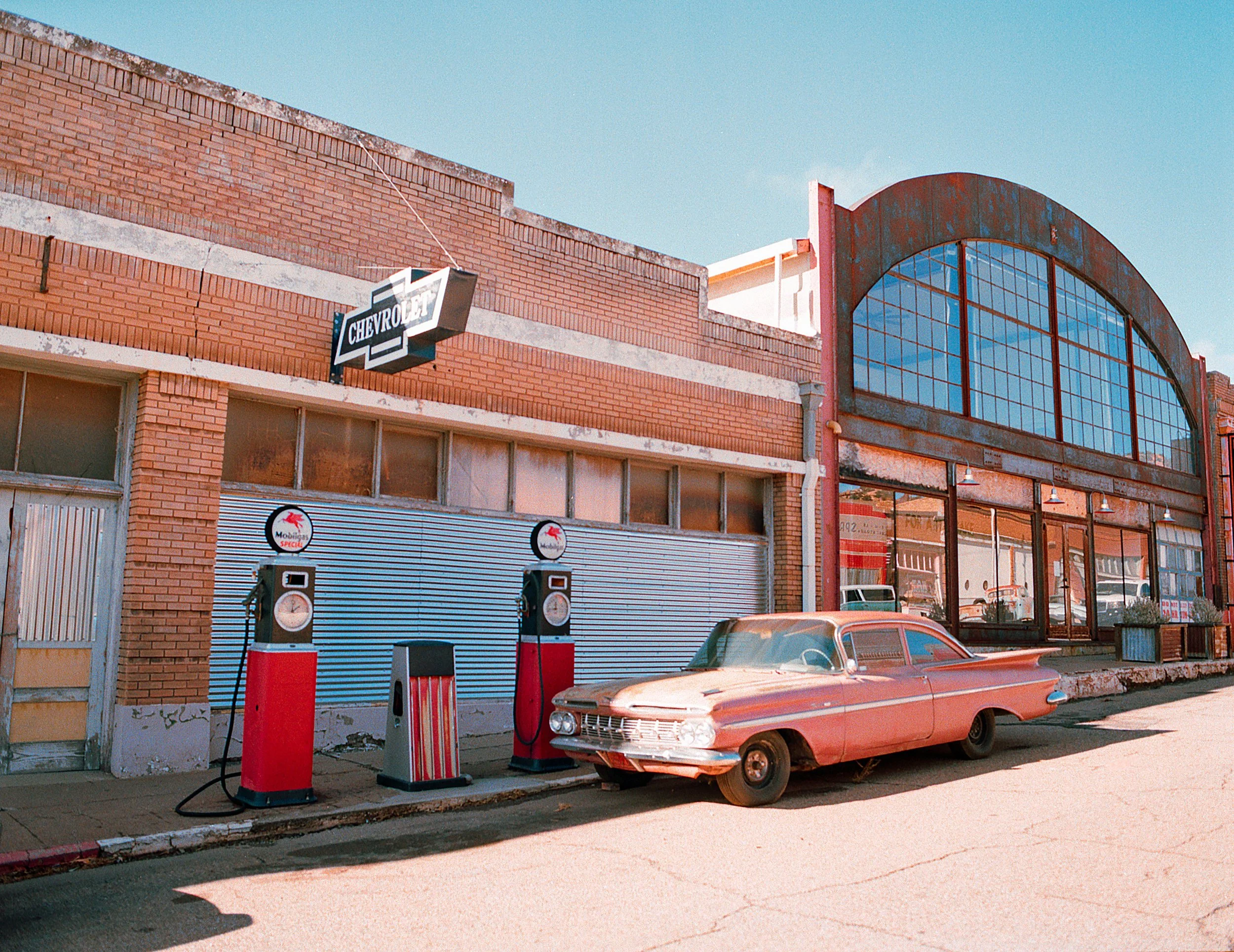 Vintage pink car parked in front of an old building with a Chevrolet sign and two old gas pumps.