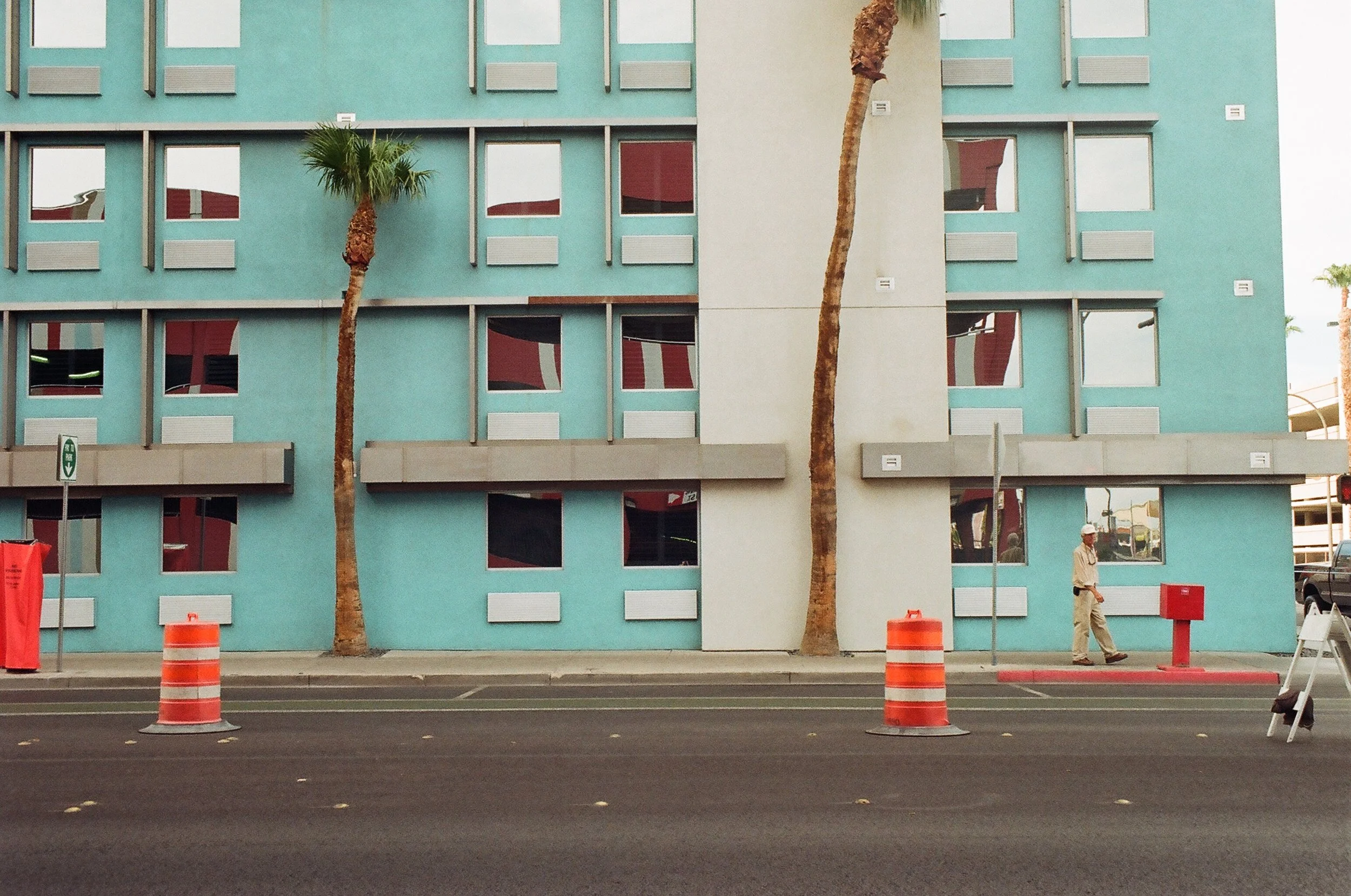 A pastel blue building with multiple windows and two tall, curved palm trees in front. There are orange traffic barrels, a red mailbox, and a man walking on the sidewalk.