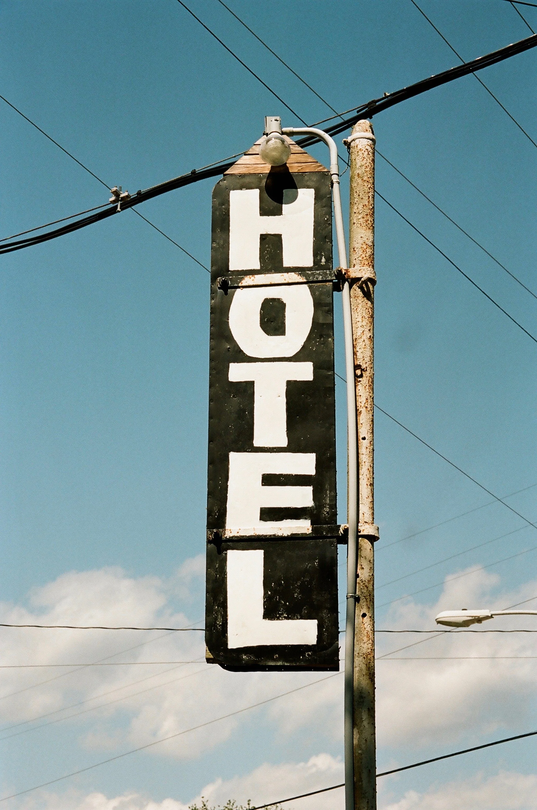 Old, weathered hotel sign hanging from a rusted pole against a blue sky with some clouds, and power lines overhead.
