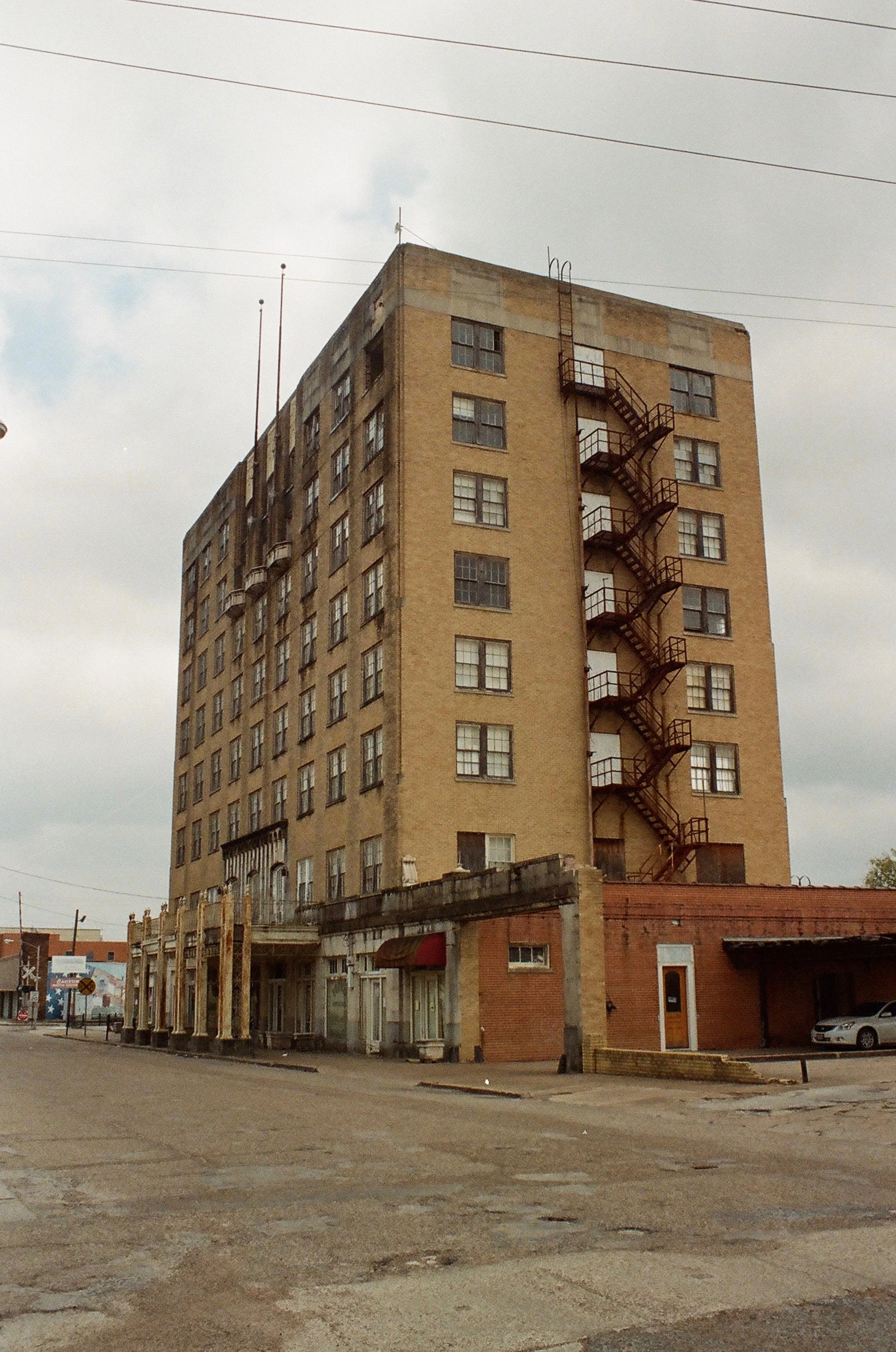 A tall, beige brick building with many windows and external fire escape stairs on the side, located on a deserted street with cloudy skies overhead.