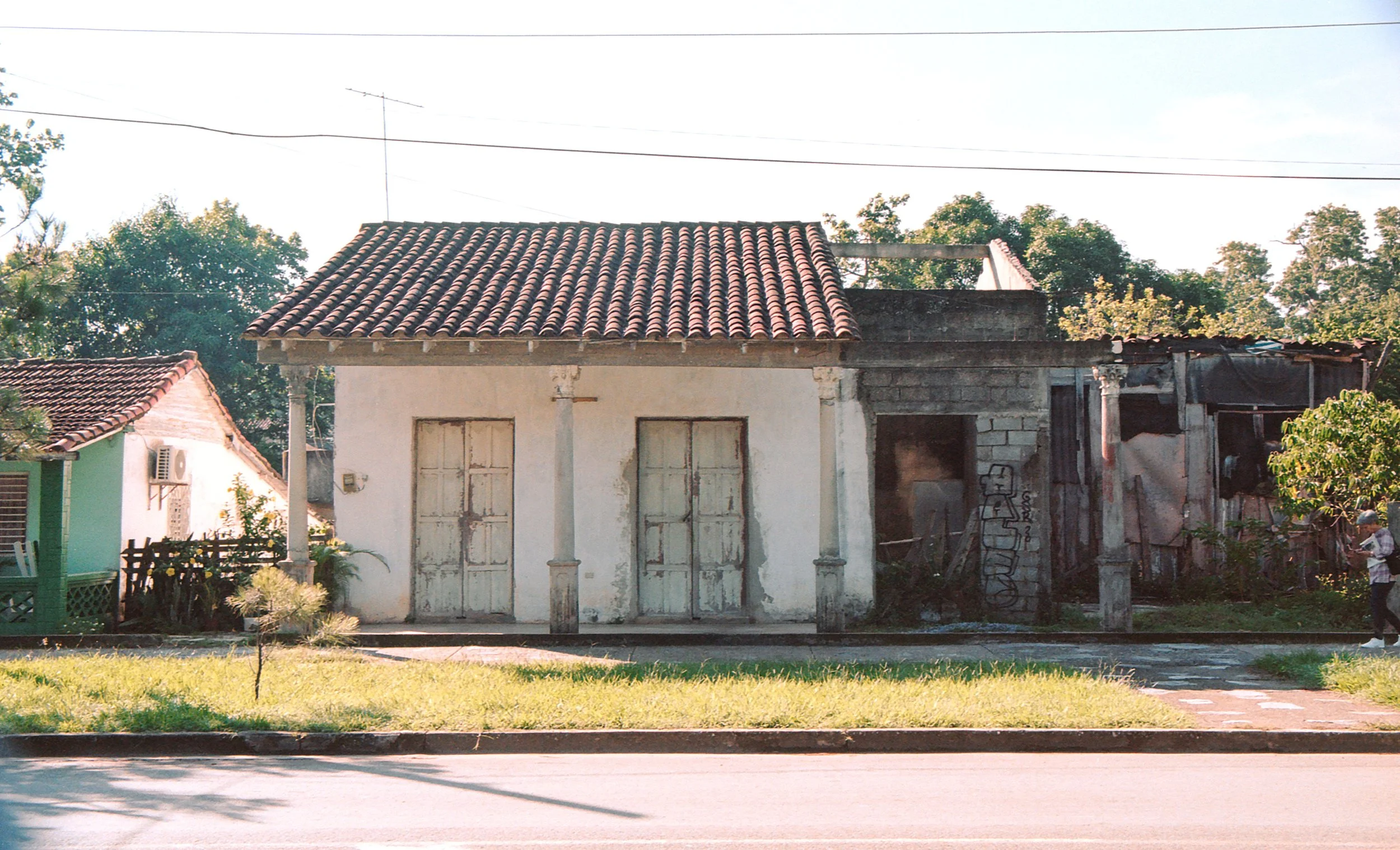 A house with a tiled roof, white walls, and two closed doors, next to a partially burned building, and a person walking on the sidewalk.