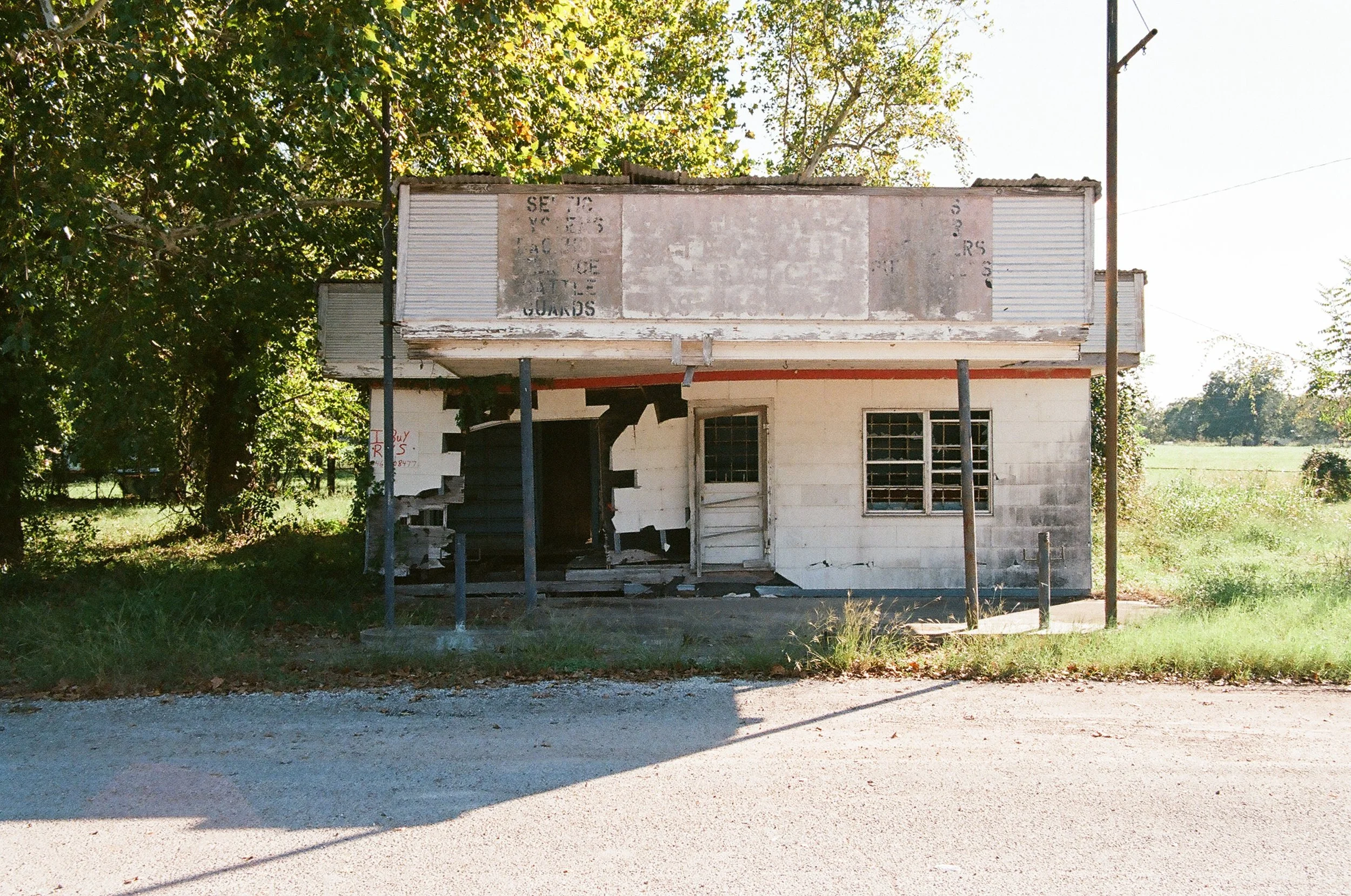 Old, abandoned small store or building with peeling paint, missing parts of the front wall, and rusted signs, surrounded by green trees and grass under a sunny sky.