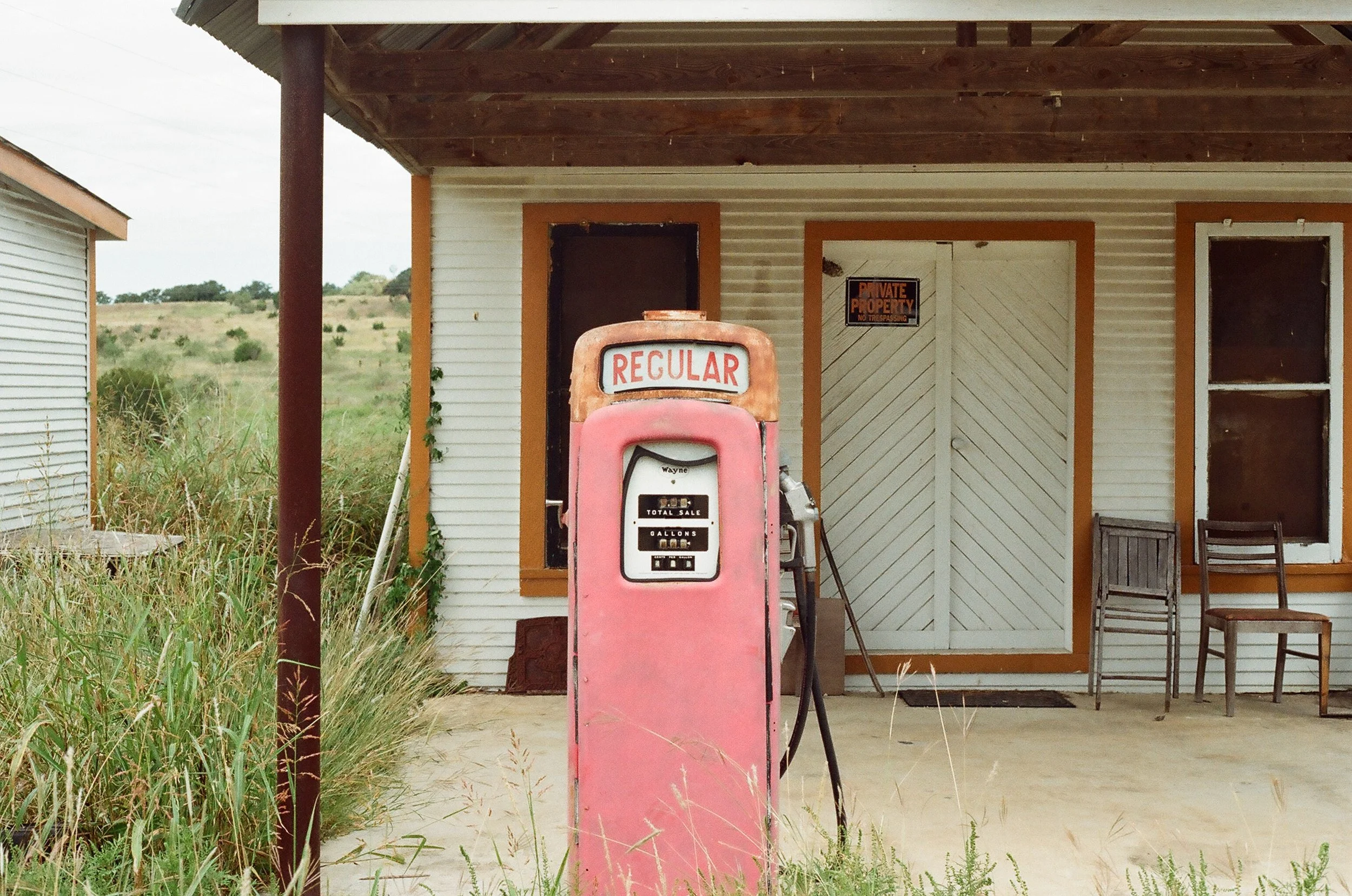 An old pink gas pump labeled 'REGULAR' in front of a rustic building with a 'PRIVATE PROPERTY' sign, empty chairs, and overgrown grass surrounding the area.