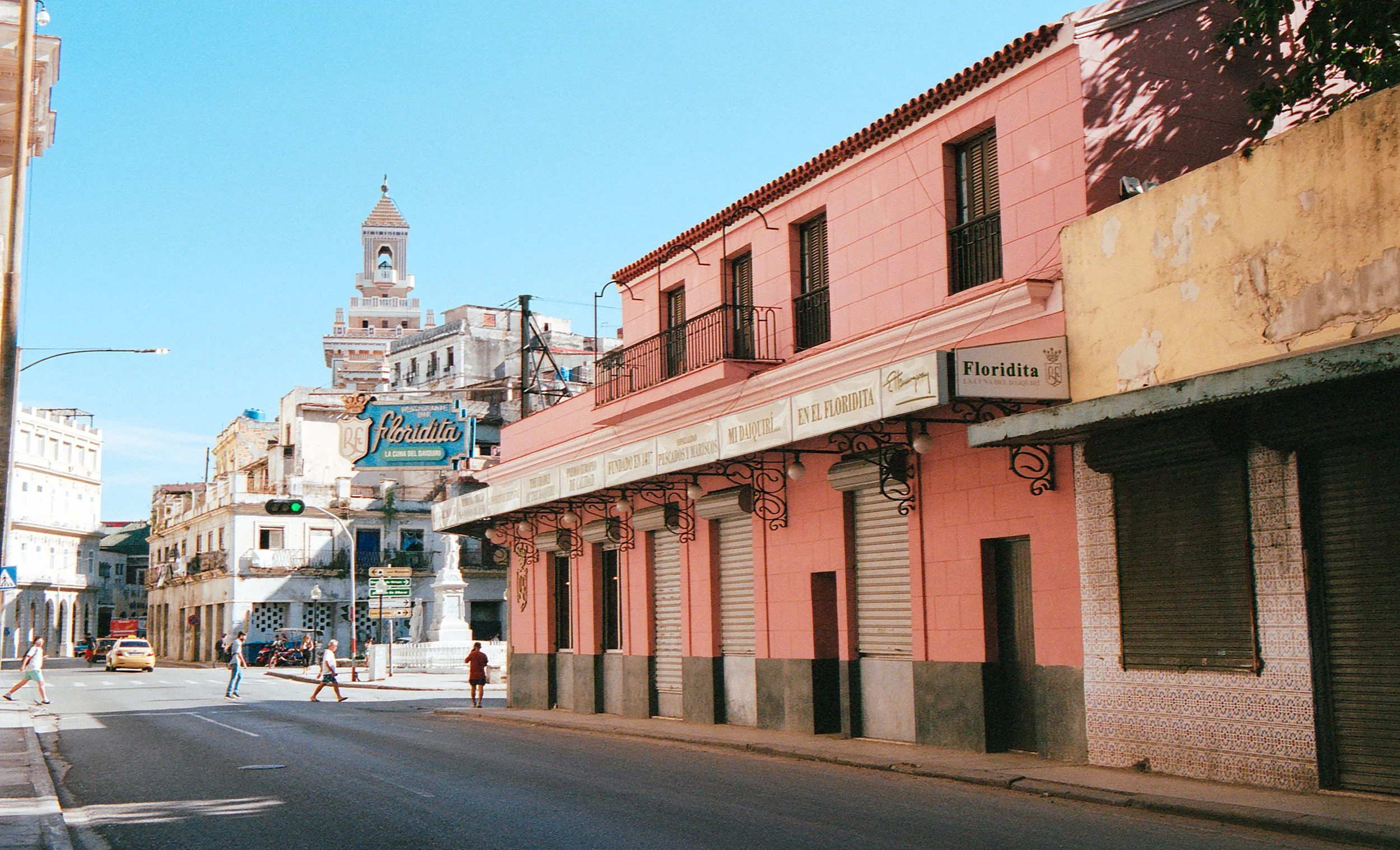 Street scene in Havana, Cuba, with pastel-colored buildings, pedestrians crossing, and a blue sky.
