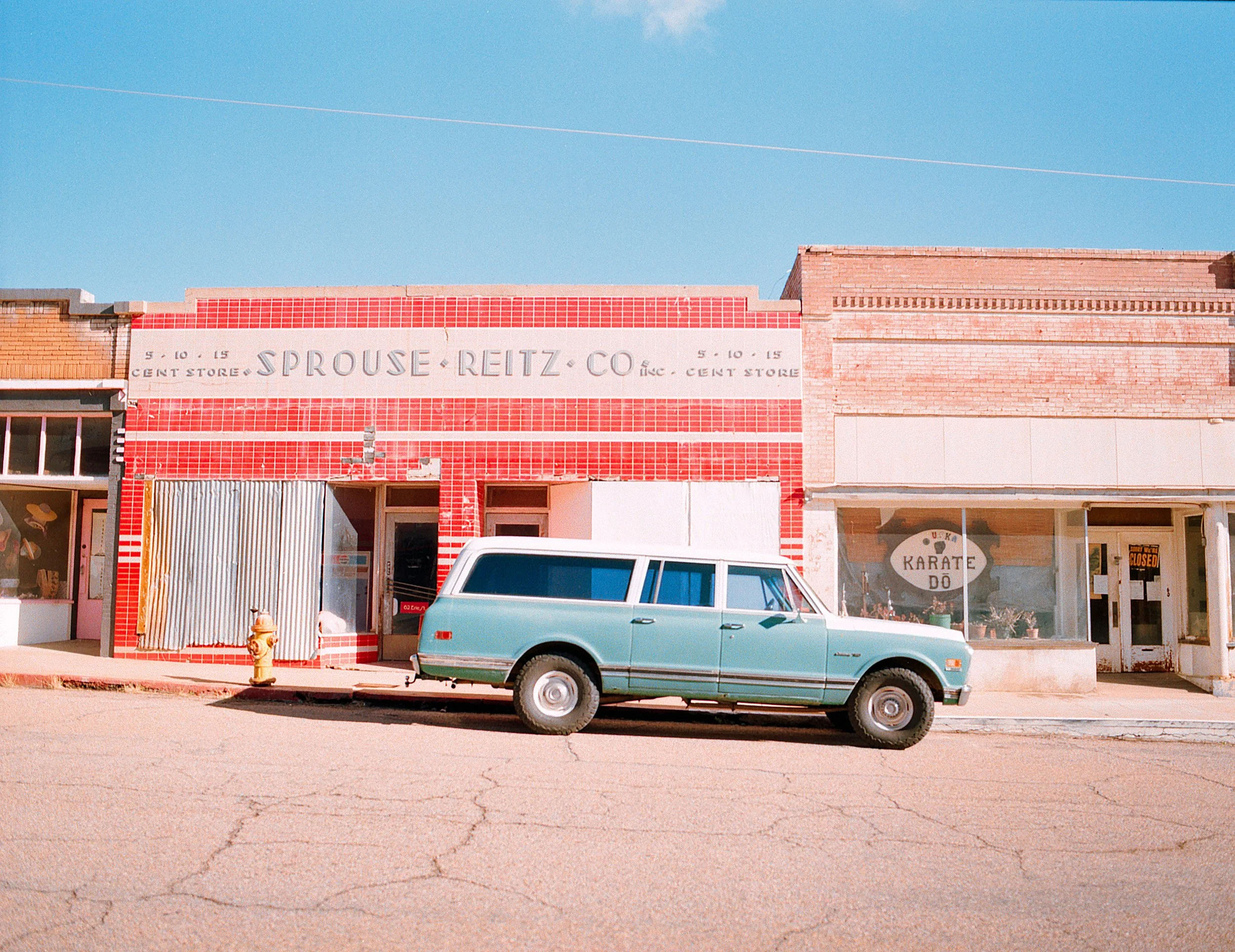 A vintage blue station wagon parked in front of small storefronts on a sunny street, with a clear blue sky overhead.
