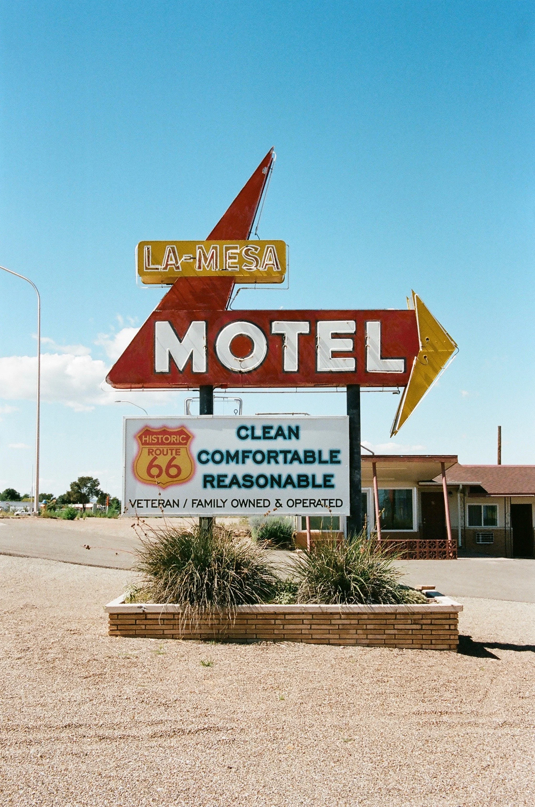 Sign for La Mesa Motel with Route 66 emblem, promoting clean, comfortable, and reasonable lodging, located in a desert area with a clear blue sky.
