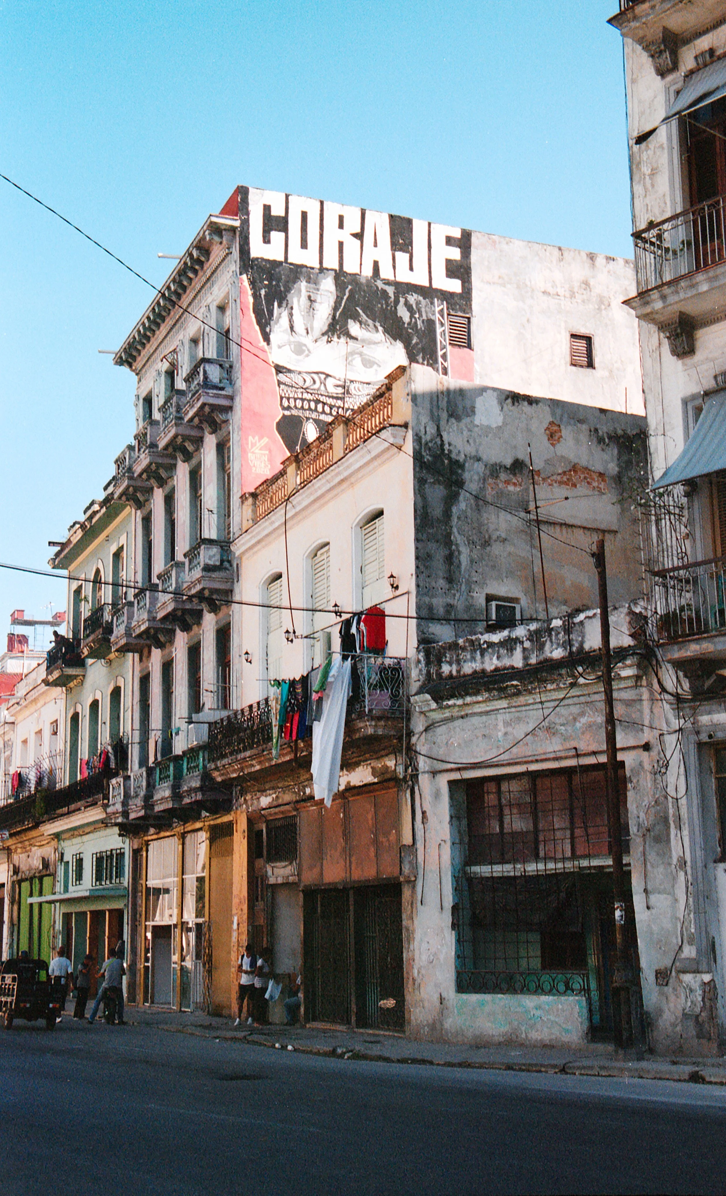 A city street with old, weathered buildings, laundry hanging from balconies, and a large mural with the word 'CORaje' painted on the top building against a clear blue sky.