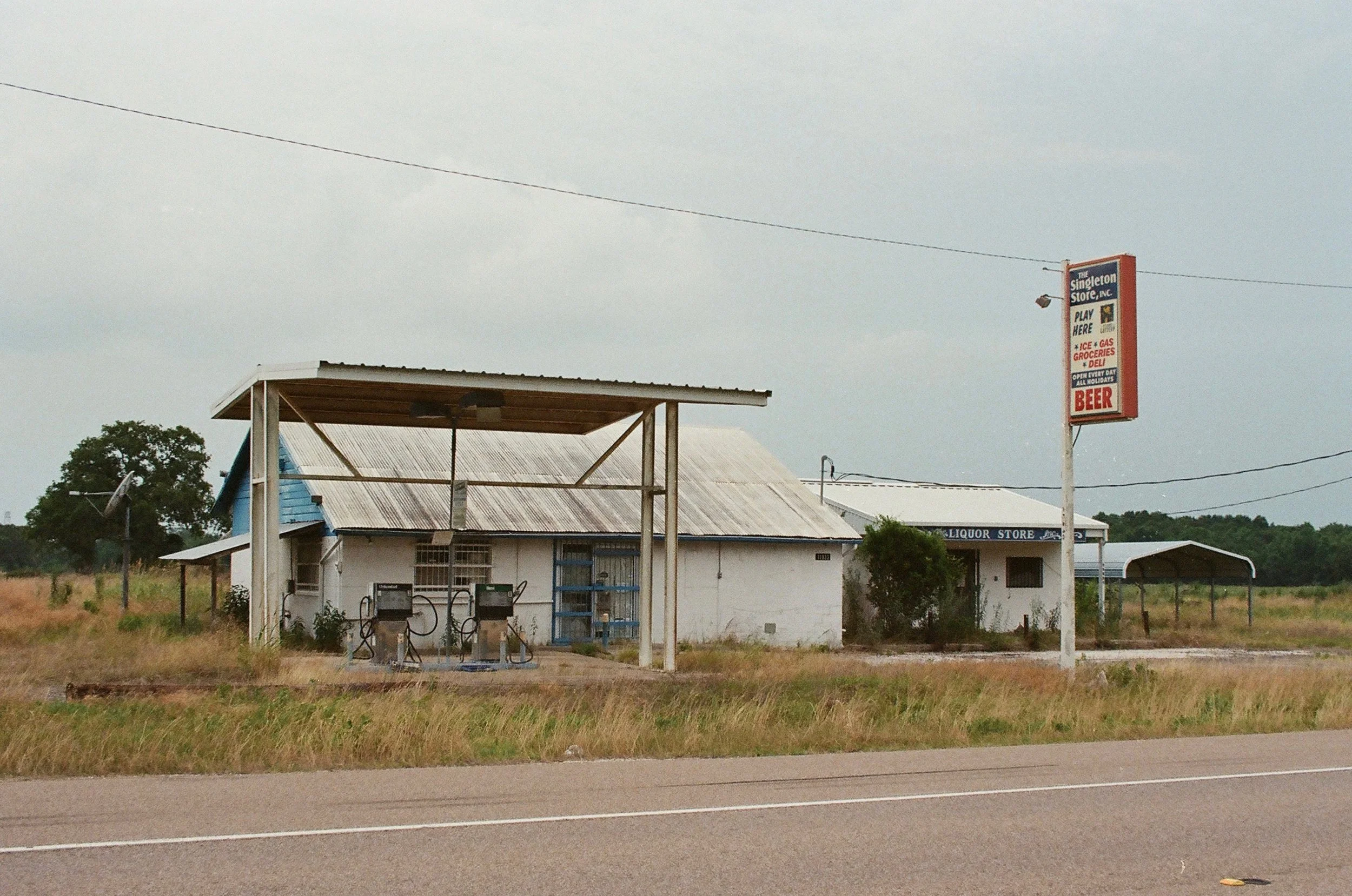 Old roadside liquor store with a weathered metal roof, two gas pumps in front, and a large sign advertising groceries, beer, and gas, set in a rural area with overgrown grass.